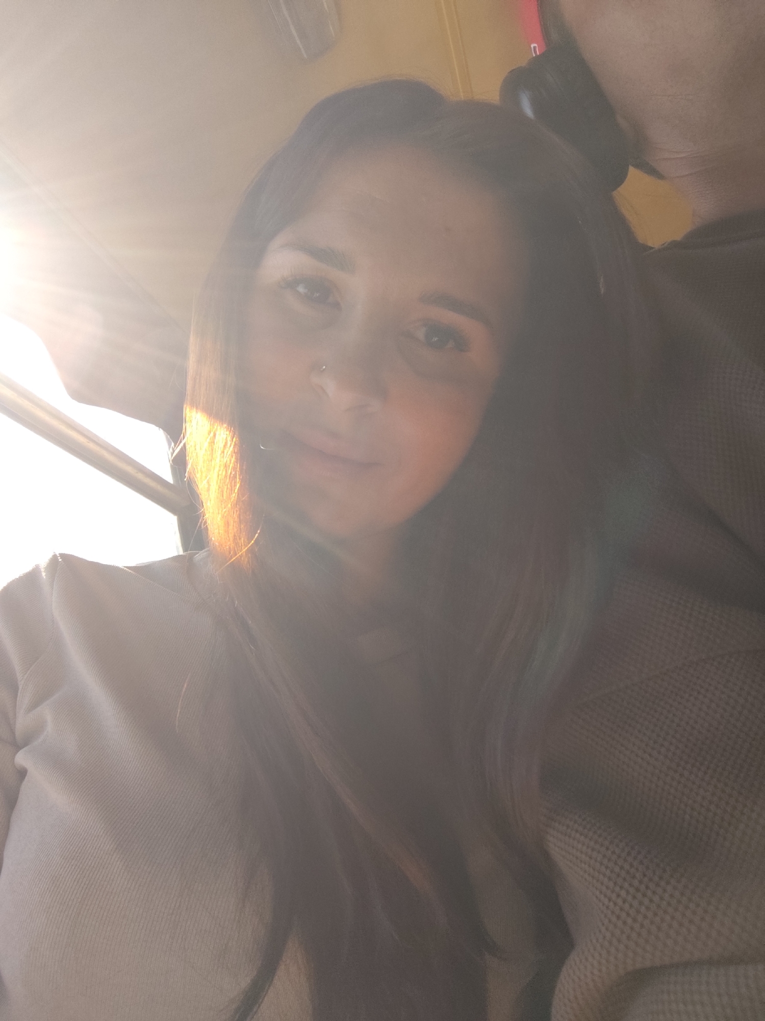 The image shows a woman with long brown hair, wearing a light-colored sweatshirt, sitting next to a person with dark hair and headphones. The woman has a nose piercing and is looking towards the camera. Sunlight is visible through a window to the left. The location is indeterminate, and there is no information to indicate that the image was taken in Chaikovsky, Russia.