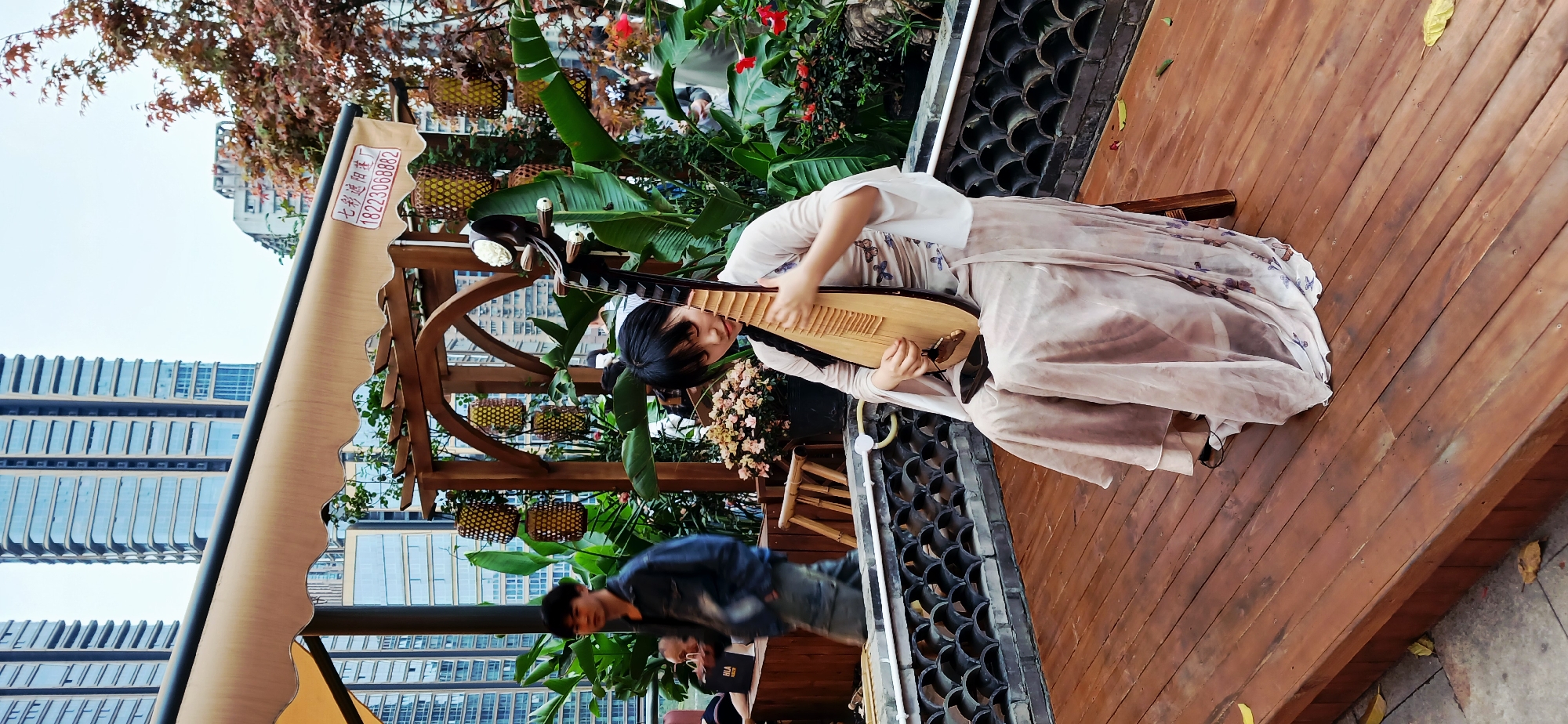 A woman in traditional Chinese dress sits on a wooden stage, playing a pipa, a Chinese lute. The scene is outdoors, with lush greenery and flowers surrounding her. A man in a denim jacket walks by in the background. Tall buildings are visible in the distance, indicating an urban setting. The scene appears to be in Yuzhong District, China.