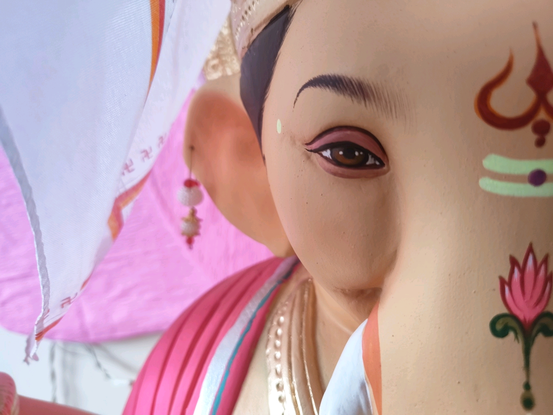 The image is a close-up shot of a Ganesh idol, focusing on its face. The idol is adorned with traditional markings and embellishments. On the forehead, a trident symbol is painted in red and orange. Below the eye, two horizontal lines in light green with a purple dot in the center are visible. A pink lotus flower with green leaves is painted on the right cheek of the idol. The idol's eye is open with brown iris and detailed black eyeliner. A small green dot is placed on the cheekbone. The idol appears to be made of clay or a similar material. In the background, a pink cloth and a white cloth with orange and yellow stripes are visible. A small beaded ornament with red and white beads hangs from the top left of the frame. The overall impression is of a devotional item, possibly prepared for a festival like Ganesh Chaturthi. Given the subject matter, this image could have been taken at a home, a temple, or a workshop in Pune, India, where the worship of Lord Ganesha is highly prevalent.