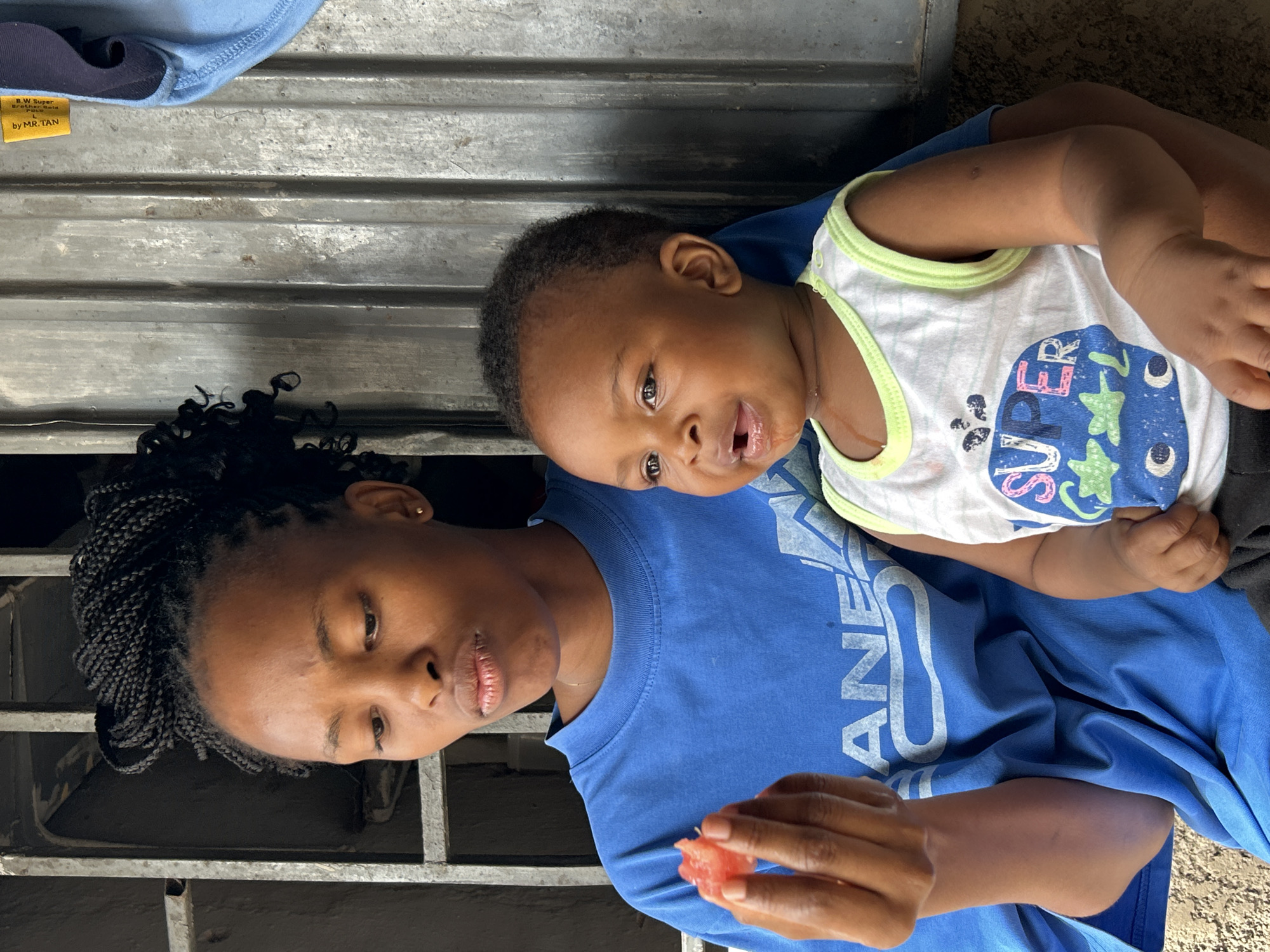 This media file is an eye-level, medium shot of a woman holding a baby. The image is oriented sideways, with the subjects appearing from left to right.
The woman, on the left, has dark skin and braided hair, with an earring visible in her left ear. She is wearing a blue t-shirt with a faint, light blue graphic design. Her eyes are closed, and her mouth is slightly open, giving the impression of a soft, possibly contented expression. Her right hand is holding a small piece of red fruit.
The baby is nestled in the woman's arms, facing slightly away from the camera. The baby has dark skin and is wearing a white onesie with a light green trim and a blue graphic design that includes stars and the word "SUPER." The baby's mouth is open, and it appears to be looking up and to the right, with a curious or engaged expression. The baby's arm is visible, resting on the woman's chest.
The background consists of a corrugated metal surface, suggesting an outdoor or industrial setting. A small yellow label with black text is visible in the upper left corner of the image, reading "B.W. Super Brother Bold L by MR.TAN." The overall lighting suggests it is daytime. The scene conveys a sense of intimacy and care between the woman and the baby.