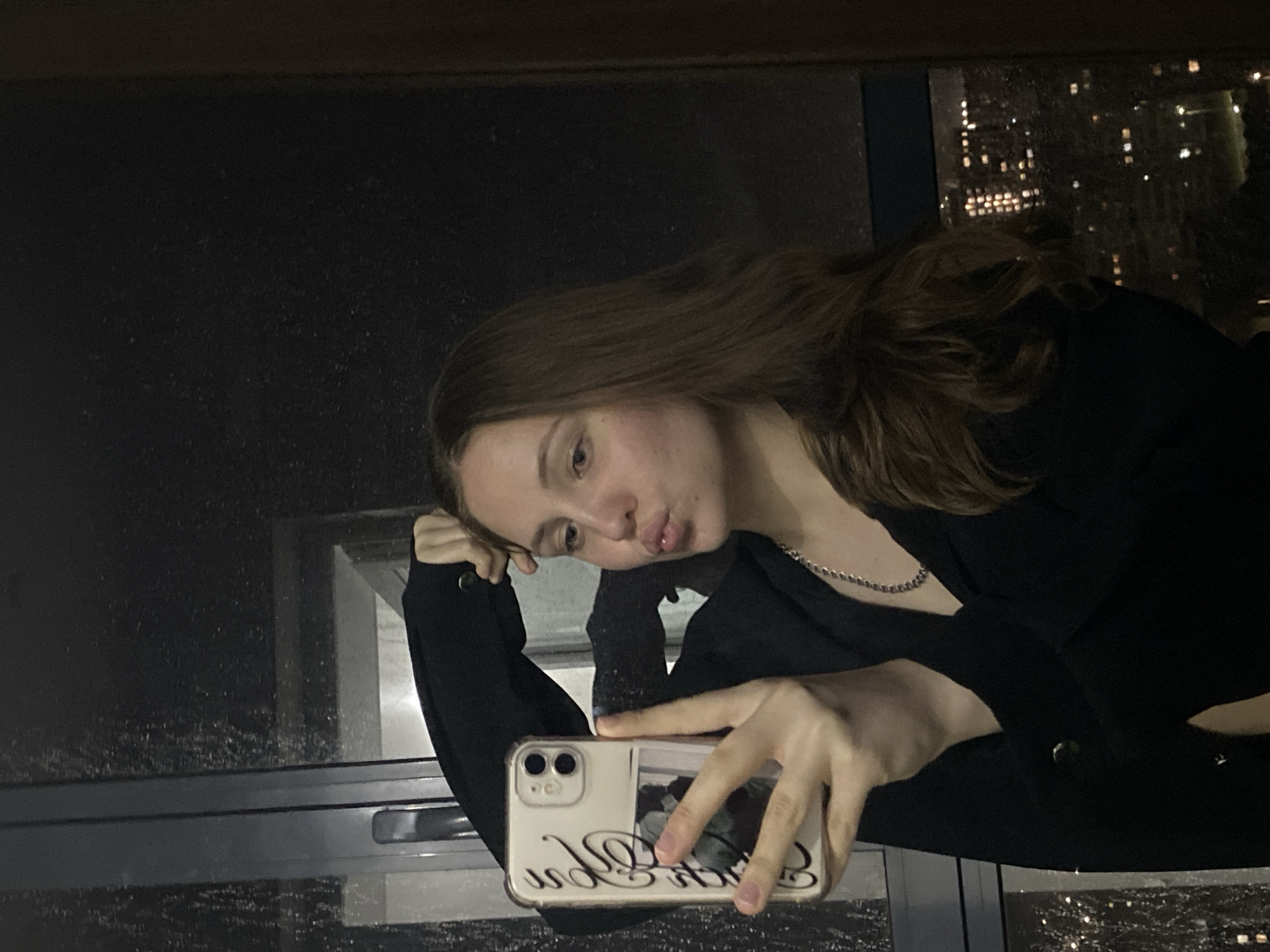 A young woman is taking a selfie indoors at night, near a large window. The window shows rain and a cityscape with lights. The woman has long brown hair and is wearing a black top or jacket. She is resting her head on her hand and making a pouting expression. She is holding a smartphone with a phone case in her other hand. The environment is dark, with city lights visible through the window. There is no information to determine if the photo was taken in Izhevsk, Russia.