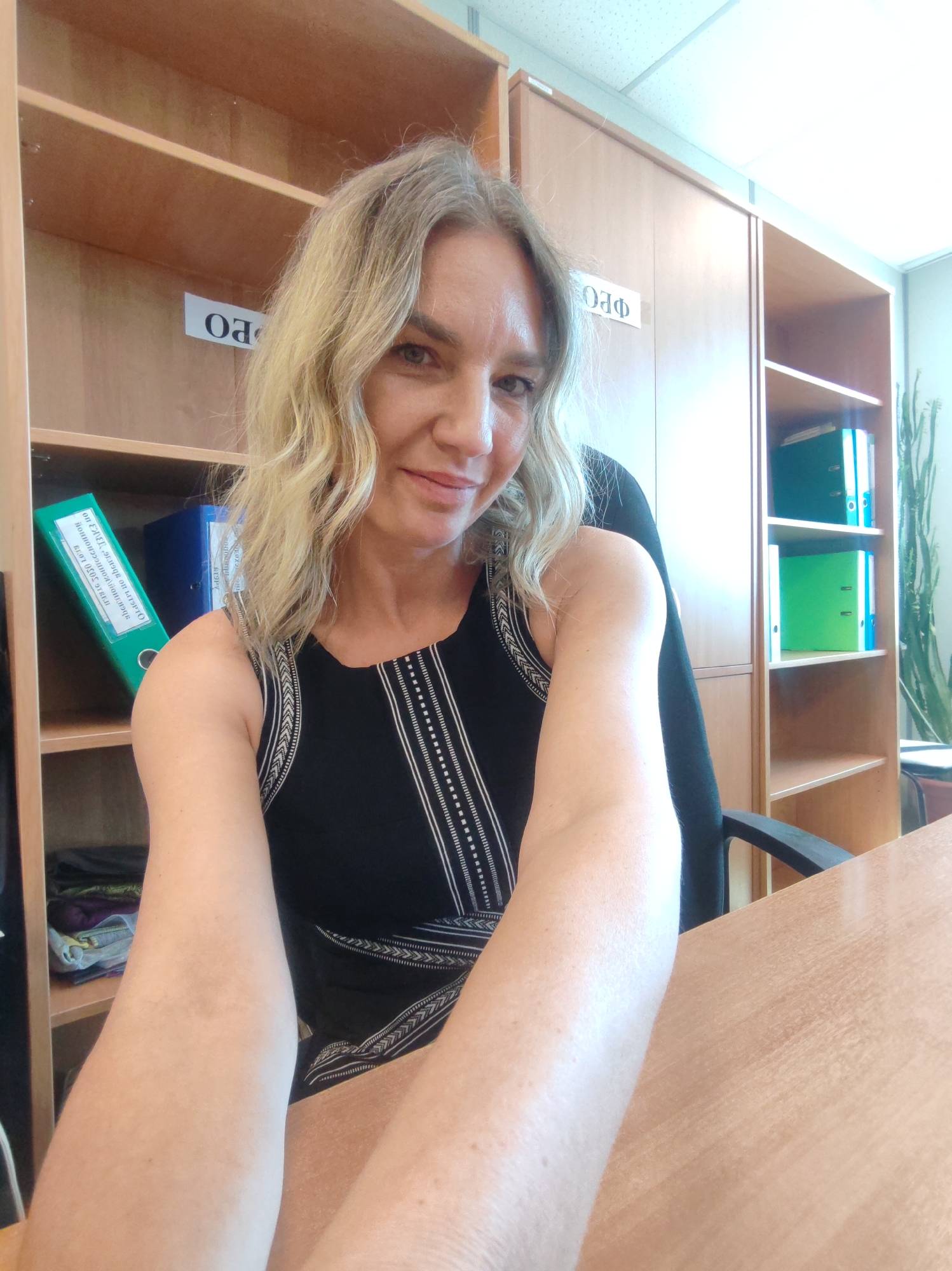 A woman with blonde curly hair and a black sleeveless dress is sitting at a wooden desk in an office setting. The office has wooden shelves filled with binders and other items. One shelf has a sign with letters, including "PO" visible. There is a tall cabinet next to the shelving unit, and a plant is visible near a window. The woman is facing the camera, and one of her arms is extended towards the viewer. OCR text on one of the binders includes mentions of "Exed," "Atorto," and a location possibly related to "3050 Long". The location in the city of Petropavlovsk-Kamchatsky, Russia is not visibly depicted in this image.