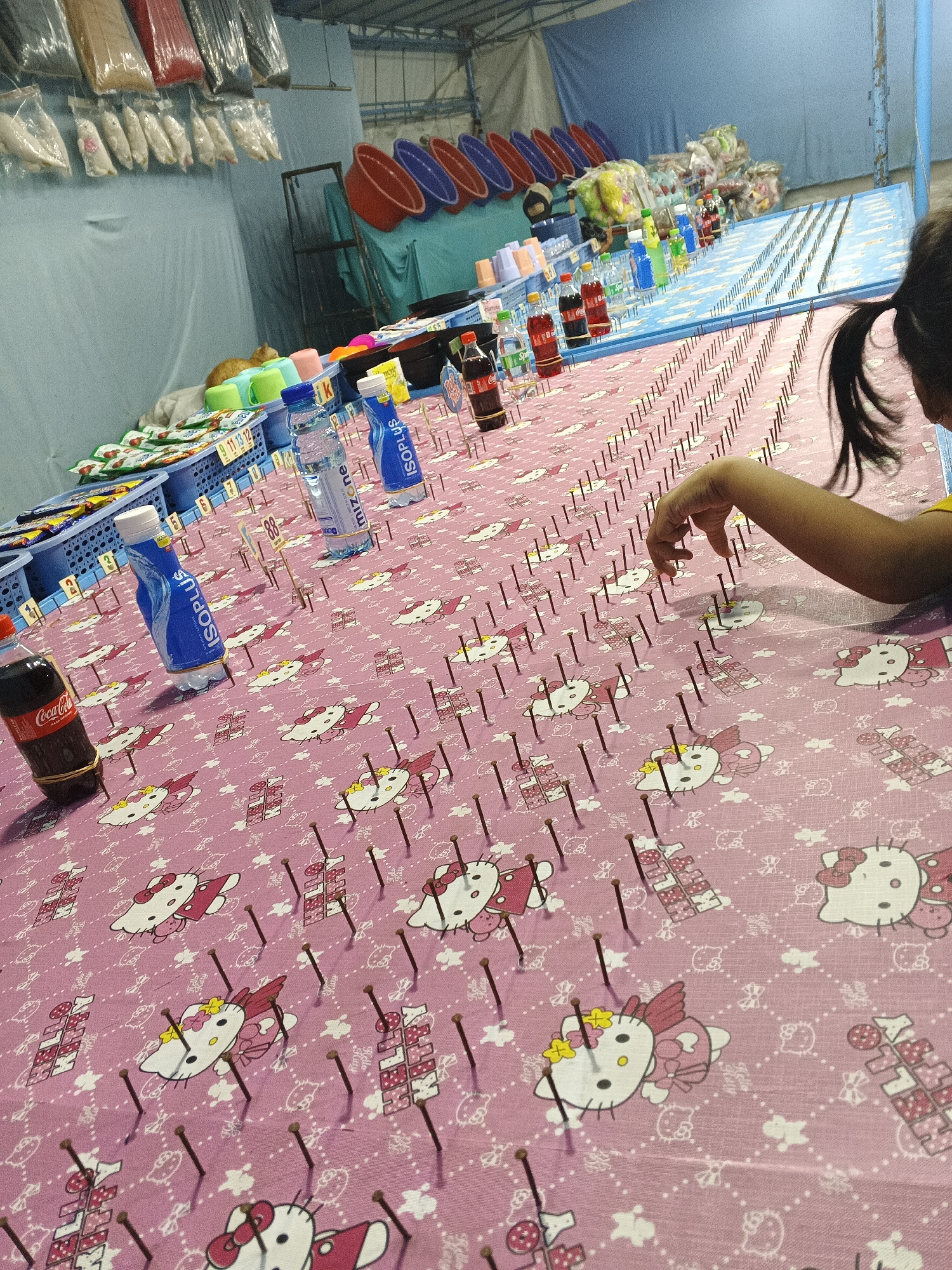 This image depicts a close-up view of a game stall, likely at a local market or festival in Sukabumi, Indonesia. The foreground is dominated by a pink cloth covered in Hello Kitty patterns, with numerous small nails stuck into it, forming a grid. A young girl, partially visible from behind, is reaching out with her right hand towards the nails, suggesting she is interacting with the game. Several bottles of beverages, including Coca-Cola and what appears to be Isoplus, are placed on the cloth, some secured with rubber bands. In the background, a variety of goods are displayed, including plastic containers, bags of merchandise, and more beverage bottles lined up on a blue surface. The overall setting appears to be an outdoor or semi-open stall.