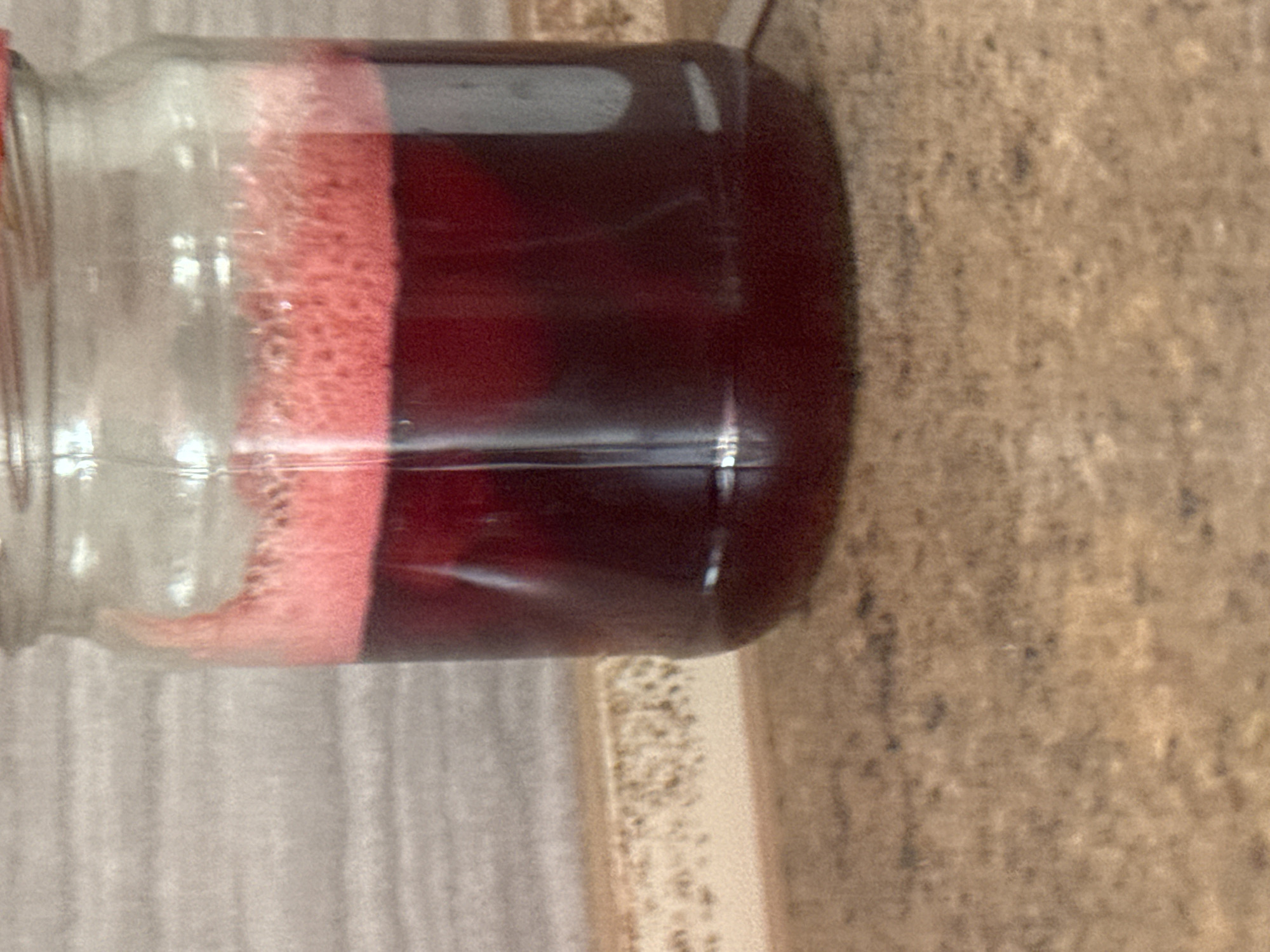 A glass jar filled with a dark red liquid and a frothy pink layer on top sits on a speckled brown countertop. Behind the jar, a grey, horizontally textured wall is visible. The image focuses on the jar and the countertop, with the background wall slightly out of focus. There are no people or active events depicted in the scene.