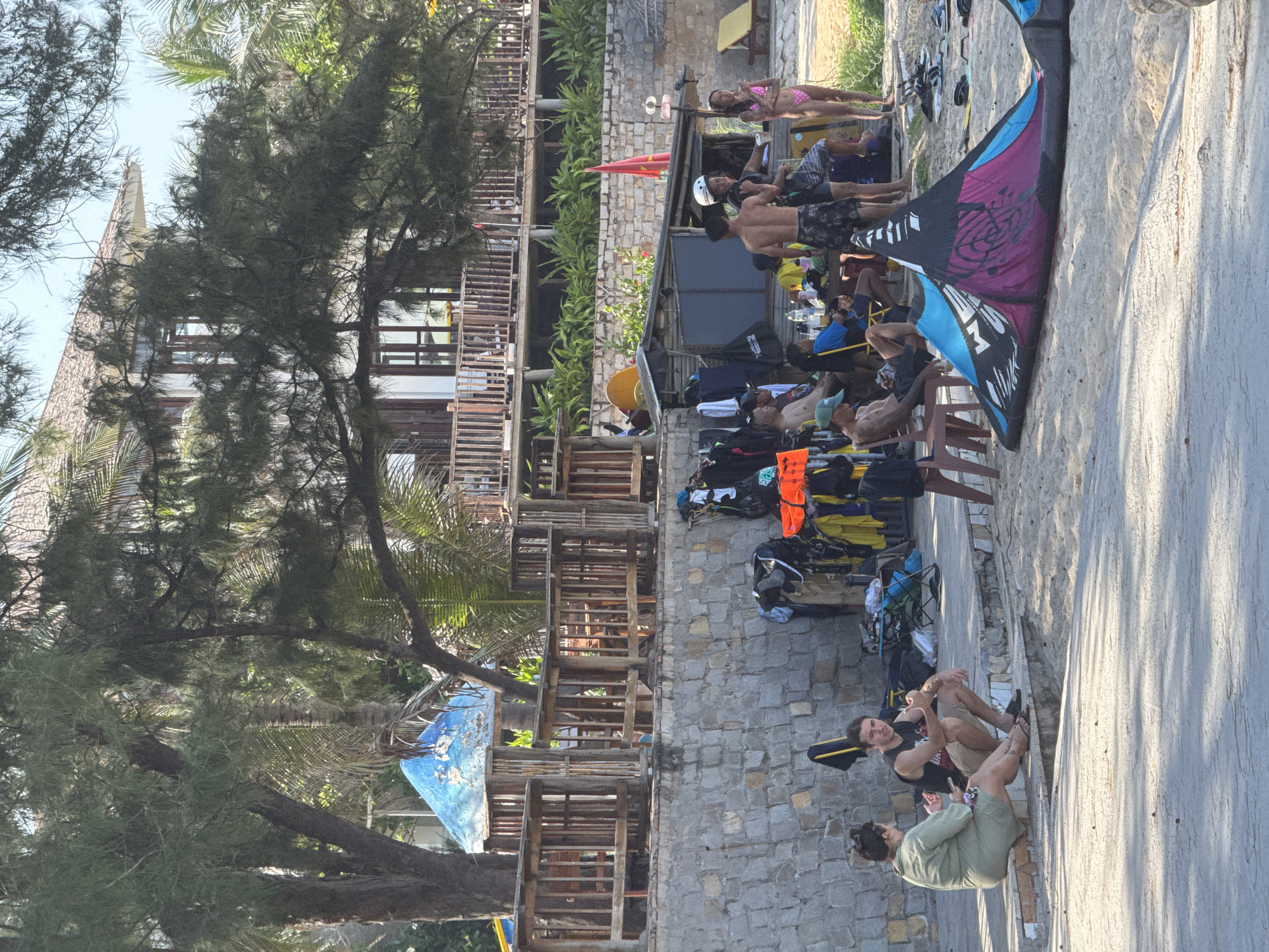 A group of people are gathered on a sandy beach in Mũi Né, Vietnam, in front of a stone wall and a multi-story building with wooden balconies. In the foreground, a colorful kite is laid out on the sand. To the left, two people are sitting on a low concrete wall; one is a woman in a green dress, and the other is a man in a black tank top and shorts. Behind them, a variety of gear, including wetsuits, life jackets, and bags, are piled near a small open-air structure. Several other individuals are standing or sitting in the background, some appearing to be engaged in water sports or related activities. Palm trees and other foliage are visible, and the overall setting suggests a recreational beach area.