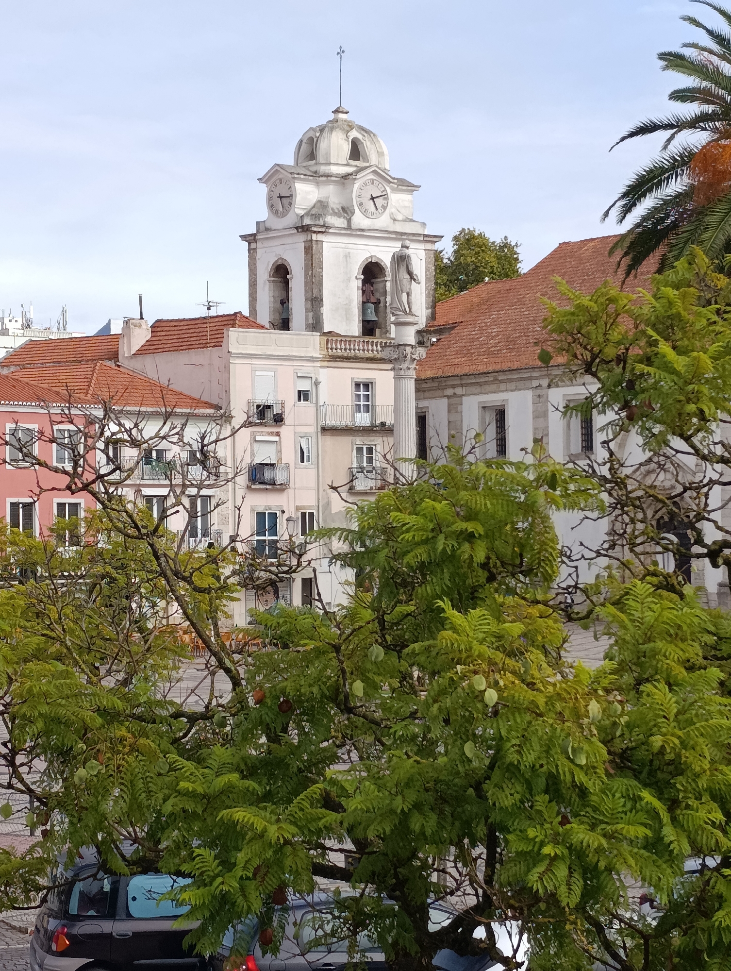 The image depicts a view of a town square in Setúbal, Portugal, featuring a prominent clock tower of a church or municipal building. The tower is white with a domed top and has two visible clock faces, each showing approximately 10:10. Below the clock faces are arched openings where bells are likely housed, and a statue stands on a pedestal in front of one of the arches.
In the foreground, lush green foliage from trees obscures parts of the scene. The leaves are small and feathery, and some pods or fruits are visible among them. Through the leaves, several buildings with red tile roofs are visible. One building is painted pink and has multiple windows and balconies. Another adjacent building is lighter in color, possibly cream or pale pink, and also features windows and balconies.
To the right of the clock tower, a portion of a building with white walls and a white stone facade is visible, with dark windows. A tall palm tree with green fronds extends into the upper right corner of the frame.
At the bottom of the image, the roof and rear of a dark-colored car are visible, parked on a cobblestone-like surface. The overall impression is a serene, daytime scene in a European town, likely a historic district.