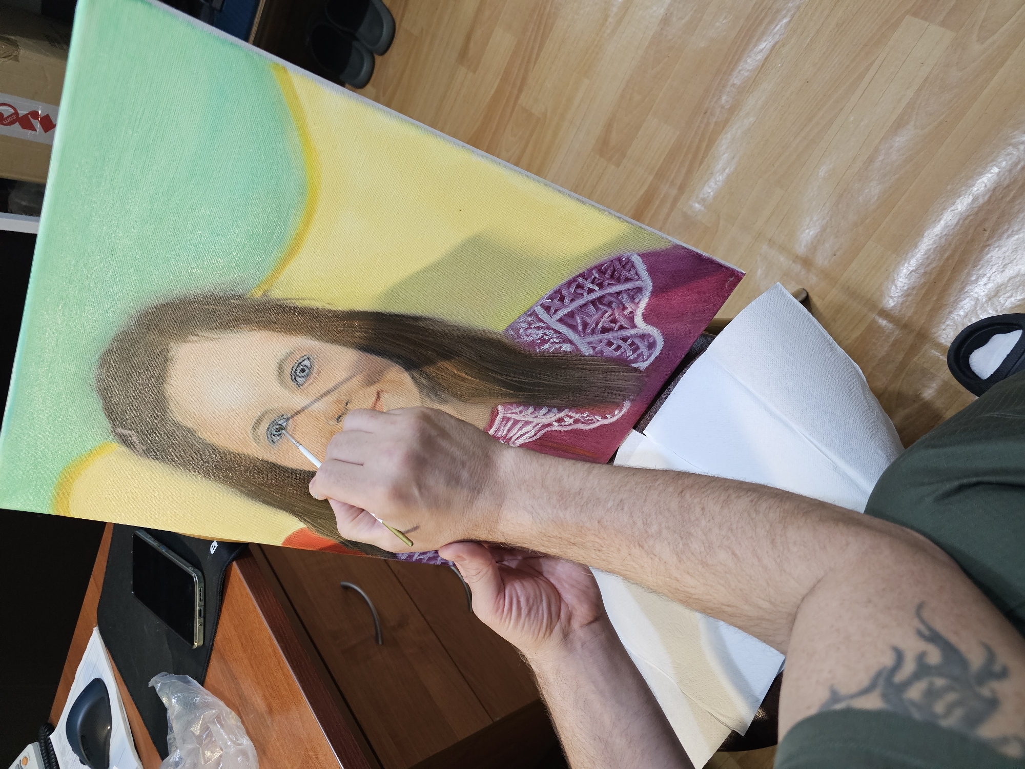A person is depicted actively painting a detailed portrait on a canvas, positioned diagonally across the frame in an indoor setting in Moscow, Russia. The canvas showcases a rendering of a young woman with long, flowing brown hair and striking blue eyes. She is adorned in a reddish-pink garment featuring intricate white patterned embellishments on the shoulder. The background of the portrait transitions from a light green on the left to a soft yellow, then a deeper yellow on the right.
The painter's arms, visibly hairy, occupy the central foreground. They are holding a slender paintbrush, meticulously adding fine details to the eye and nose region of the portrait, demonstrating a focused and precise technique. A dark, tribal-style tattoo is visible on the upper part of the painter's right arm. White paper towels are placed beneath their painting arm.
The immediate surroundings suggest a workspace, possibly a home studio. A wooden desk or drawer unit is visible to the lower left, holding a black smartphone resting on a dark mouse pad, a black computer mouse, and a crumpled white plastic bag. The floor, visible in the background, is made of polished wooden planks that reflect light. A pair of black slippers can be seen near the top right of the floor. In the far left background, a cardboard box with a partially visible red and white sticker displaying "com" is present. The scene is well-lit, indicating daytime.