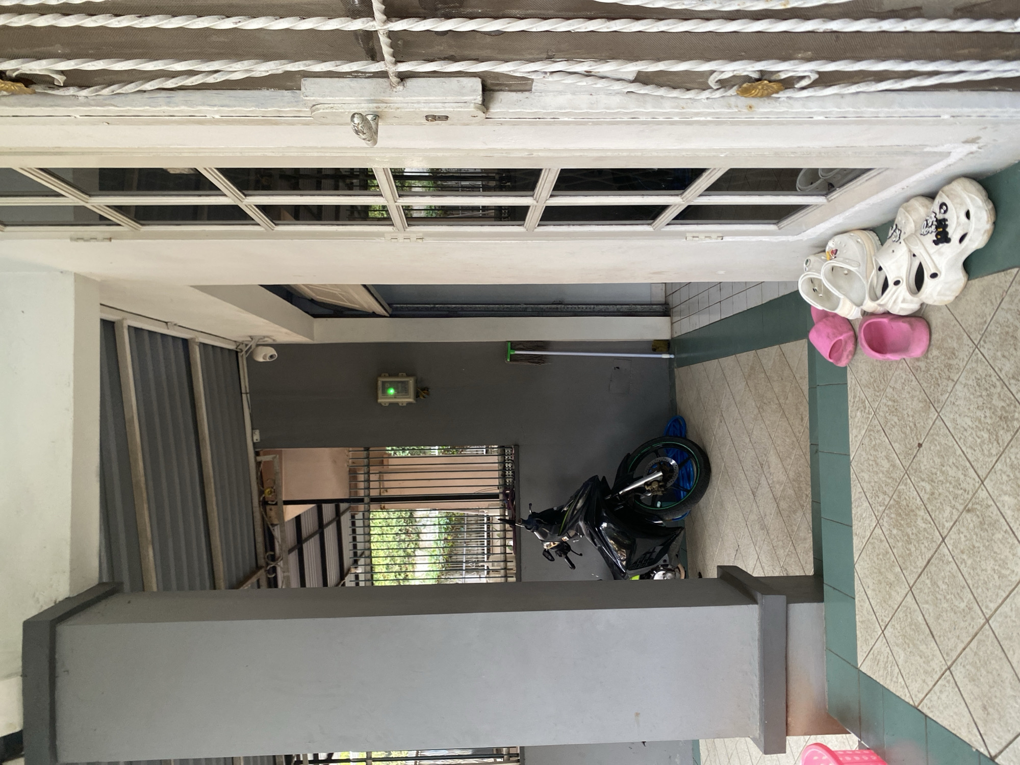The image depicts a residential exterior, likely in Dukuh, Indonesia, featuring a covered porch area. A dark-colored motorcycle is parked on the tiled floor, partially obscured by a thick grey pillar. To the right of the pillar, a white door with multiple glass panes and a decorative metal grille is visible. A set of white clogs and pink slippers are placed on the tiled floor near the door. Further into the background, a green light is illuminated on a wall-mounted electrical box, and a mop with a green handle stands upright. A security camera is mounted on the wall. The environment suggests a domestic setting with common household items.