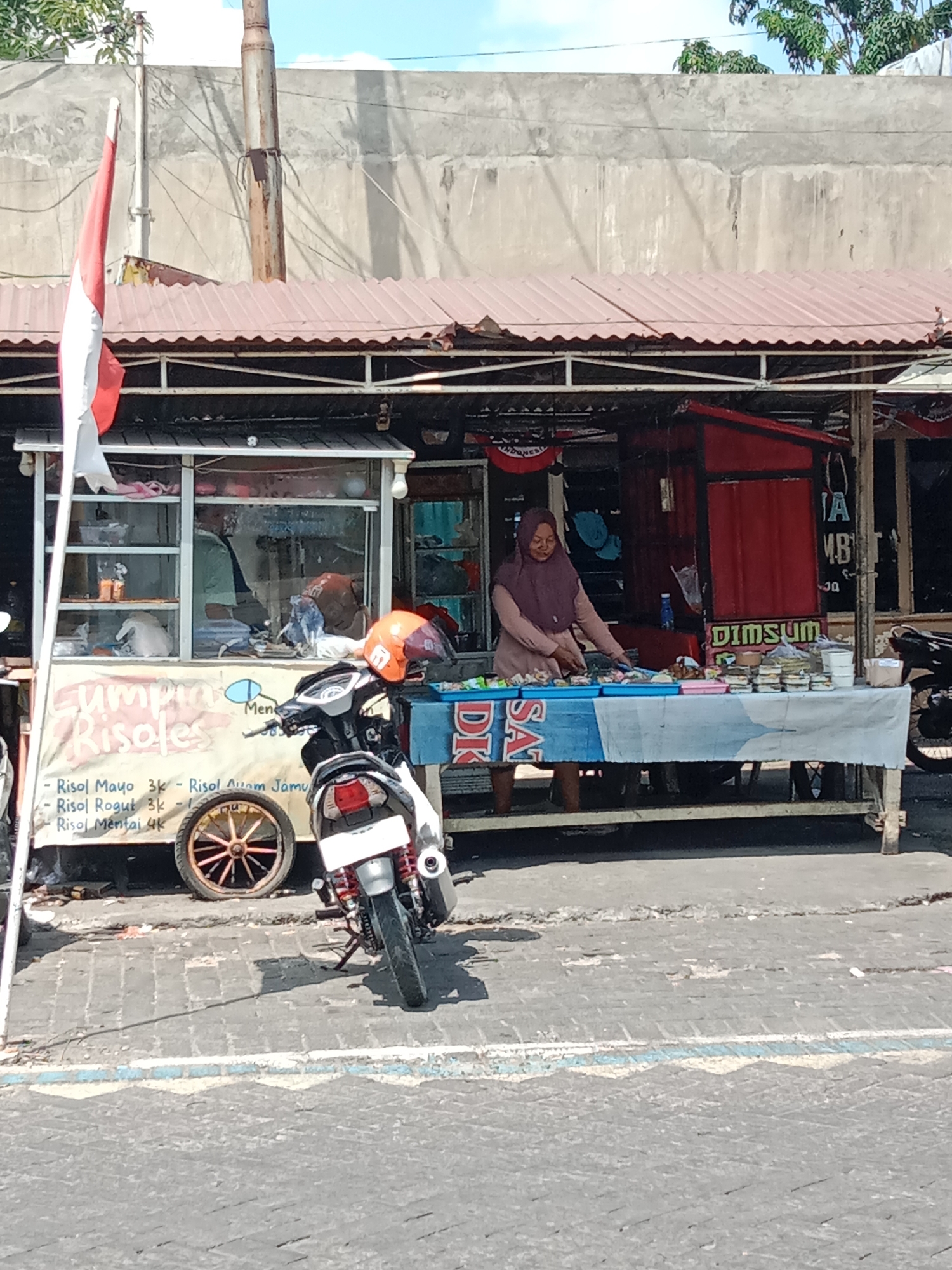 The image depicts a street food stall in Surabaya, Indonesia. The stall, situated outdoors, features a covered counter displaying various food items. A woman in a headscarf is seen attending to the food. Adjacent to the stall, a motorbike is parked, and a cart is also visible. The background includes a textured wall and trees. The overall scene suggests a typical street-side commercial activity in a city environment.