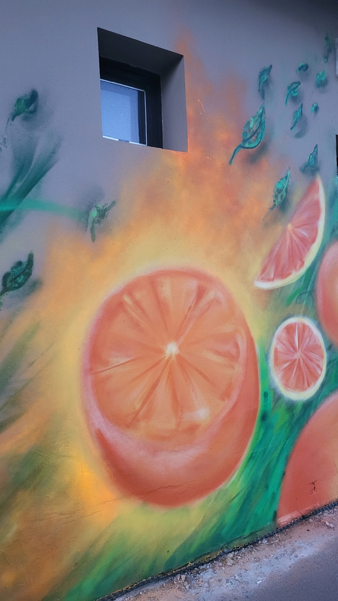 This image depicts a mural on the exterior wall of a building in Prešov, Slovakia. The mural features stylized depictions of oranges, with a large cross-section of an orange dominating the center of the frame. There are also slices of oranges and green leaf-like shapes scattered across the wall, rendered in a spray-painted style with vibrant orange, yellow, and green hues. A small, dark-framed window is visible in the upper left portion of the wall. The wall itself is painted a neutral gray. The lower edge of the image shows a sidewalk or ground area with gravel and a dark metal trim.