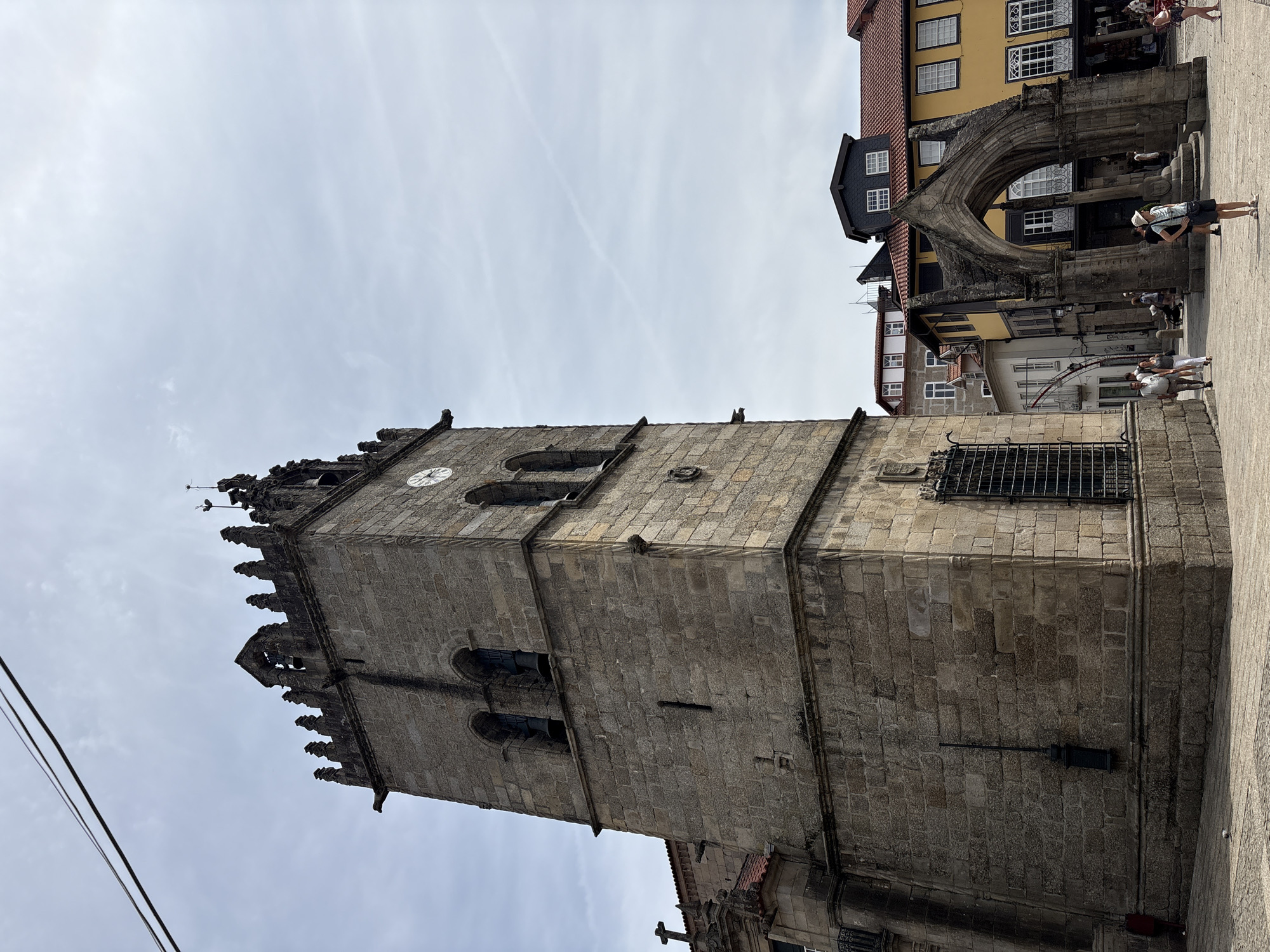 This image captures a view of the historic center of Guimarães, Portugal, specifically focusing on the bell tower of the Church of Nossa Senhora da Oliveira. The stone bell tower, with its Gothic architectural elements including battlements and arched windows housing bells, dominates the left side of the frame. A clock face is visible on one side of the tower. To the right of the tower, an ancient stone archway stands in a public square, framing a view of colorful buildings with traditional Portuguese tile roofs. Several people are walking through the square, some pausing near the archway, while others are visible in the background among the shops and cafes. The sky is overcast with a few wispy clouds. The overall scene depicts a sunny day in a well-preserved historic urban setting.