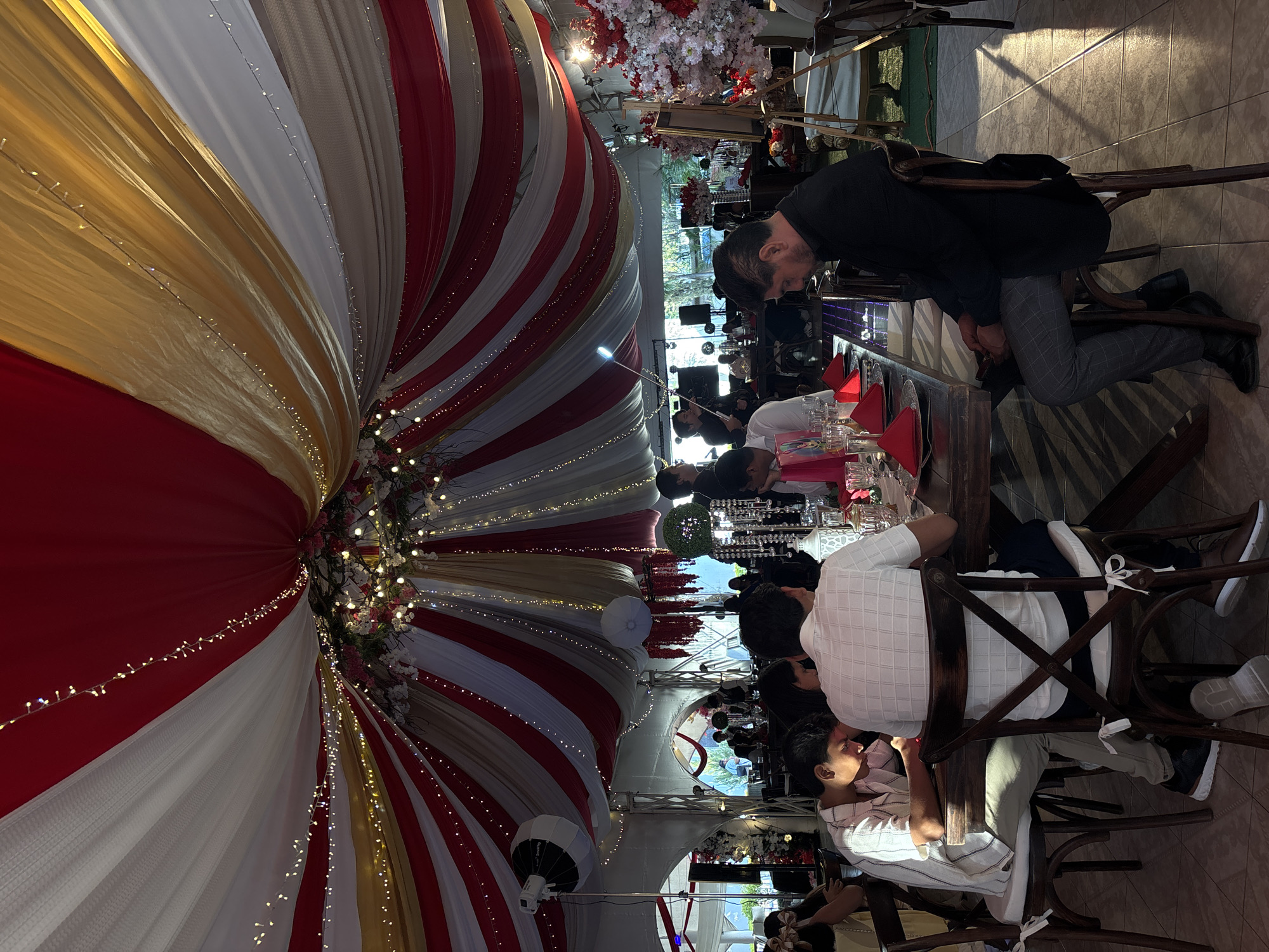 The image depicts an indoor event space in Chilpancingo, Mexico. A large, striped canopy in red, white, and gold fabric drapes overhead, illuminated by string lights. Below, people are seated at tables set with glassware and red napkins, appearing to be at a meal. An adult in a white and brown plaid shirt sits at the table, next to another individual in a striped shirt. The floor has a tiled pattern.