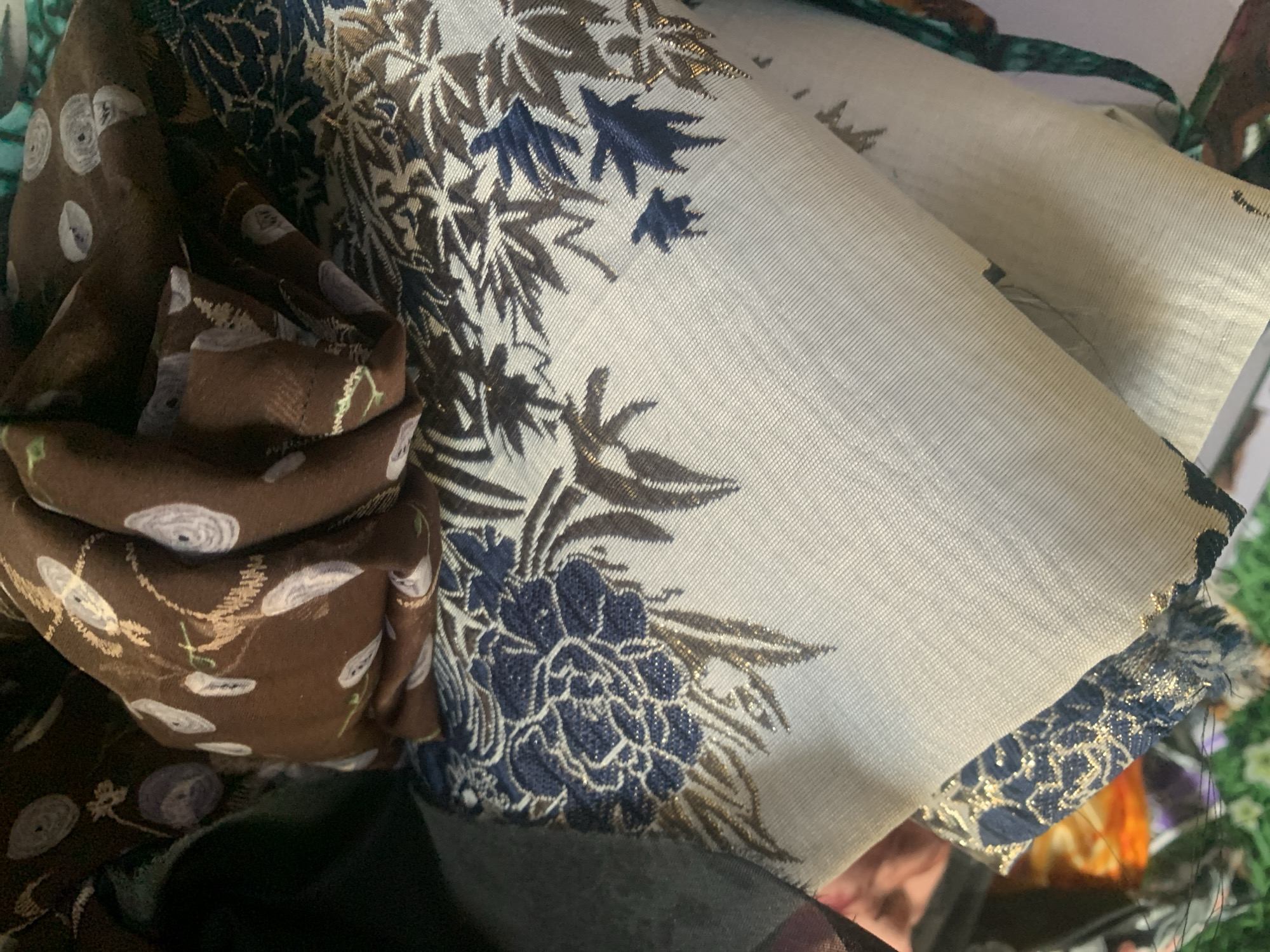 The image shows a close-up view of several pieces of fabric, stacked and folded. The fabrics have intricate patterns, including floral designs in shades of brown and blue, as well as gold metallic threads. One fabric is a deep brown with lighter brown and white circular and floral motifs. Another fabric is a lighter color, possibly cream or light grey, with detailed blue and brown floral patterns and accents of gold. The bottom edge of this fabric appears to have a textured blue and gold trim. The fabrics are arranged in a way that suggests they are either being stored or displayed, possibly in a market or shop. There is no discernible context of Jauro Yinu, Nigeria within the image itself.