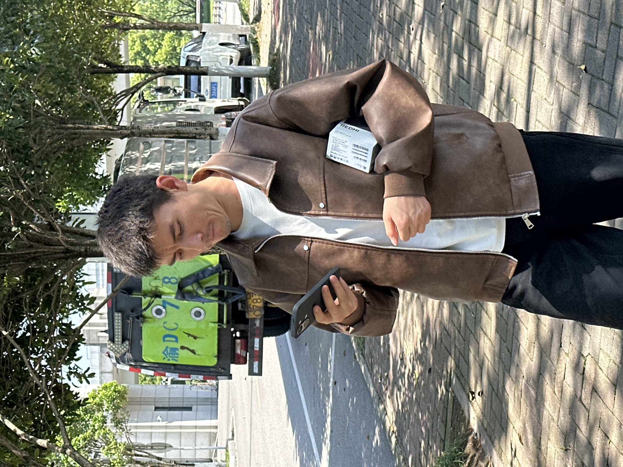 A man stands outside in Jiangjin, China, holding a phone. He is wearing a brown jacket, white shirt, and black pants. The background includes a truck with the license plate渝DC7878. The surface he is standing on is paved with interlocking bricks. The environment is sunny with shadows cast on the ground.