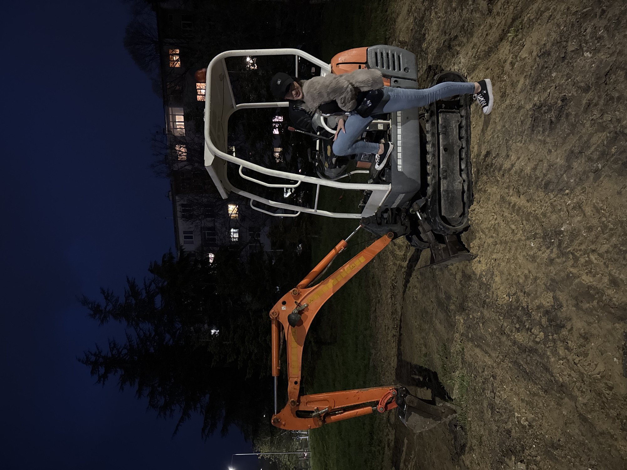 A young woman is shown playfully posing on a small, orange and grey excavator at night in Varna, Bulgaria. The woman, who has dark hair and is wearing a black cap, a dark jacket, a fluffy grey vest, and blue jeans, sits in the operator's seat of the excavator, smiling towards the camera. Her left foot, wearing a black sneaker with a white sole, rests on the excavator's rubber track.
The excavator features an open white canopy or roll cage, an orange hydraulic arm with a bucket, and grey engine housing. The ground beneath and around the machinery is dark, muddy, and uneven, suggesting a construction site or unpaved area. In the background, under a dark blue night sky, large, silhouetted trees stand in front of multi-story buildings, some with illuminated windows, indicating an urban or suburban setting. The scene is lit by ambient artificial light sources, casting shadows and highlighting parts of the excavator. The overall atmosphere is casual and lighthearted. No discernible text is visible on the excavator or in the background.
