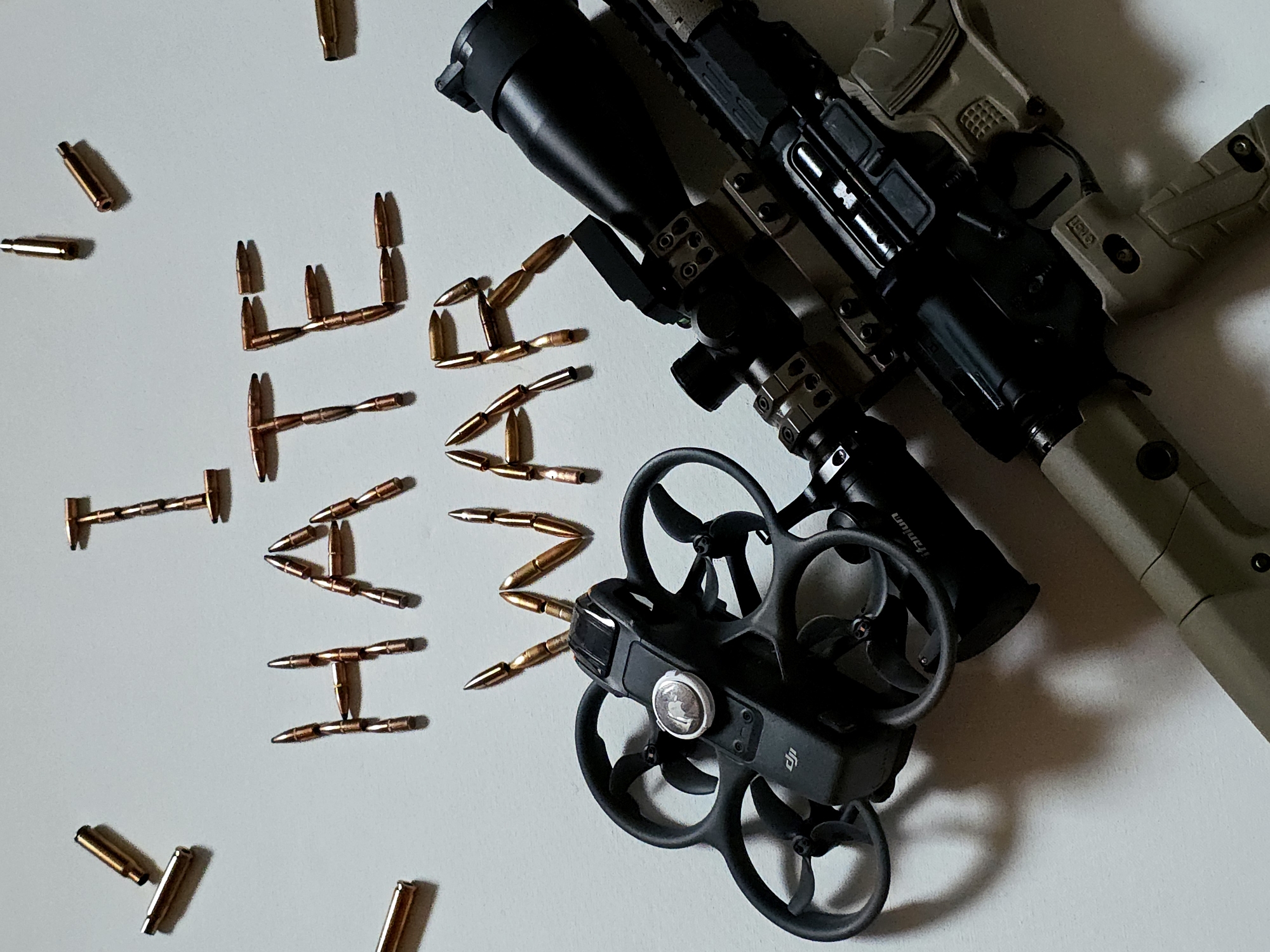 An overhead, flat-lay photograph displays a controversial arrangement of objects on a light-colored surface. In the center, numerous copper-colored rifle bullets are meticulously arranged to spell out the word "HATE."
To the right of this arrangement, a modern sporting rifle, likely an AR-style firearm, is visible. The rifle features a black upper receiver, a tan or flat dark earth (FDE) lower receiver, pistol grip, and stock. It is equipped with a black scope, which has "titanium" visible on its barrel. Below the rifle, a black DJI drone, possibly a DJI Avata or FPV model, is positioned. The drone has distinct propeller guards, and the "DJI" logo is visible on its body. A white Apple AirTag is attached to the top of the drone. Additional loose bullets are scattered around the main arrangement.
The scene is brightly lit, suggesting an indoor setting under artificial or diffused light, creating soft shadows. The overall composition appears to be a staged symbolic statement, using objects often associated with conflict, surveillance, and weaponry to convey a powerful and provocative message.