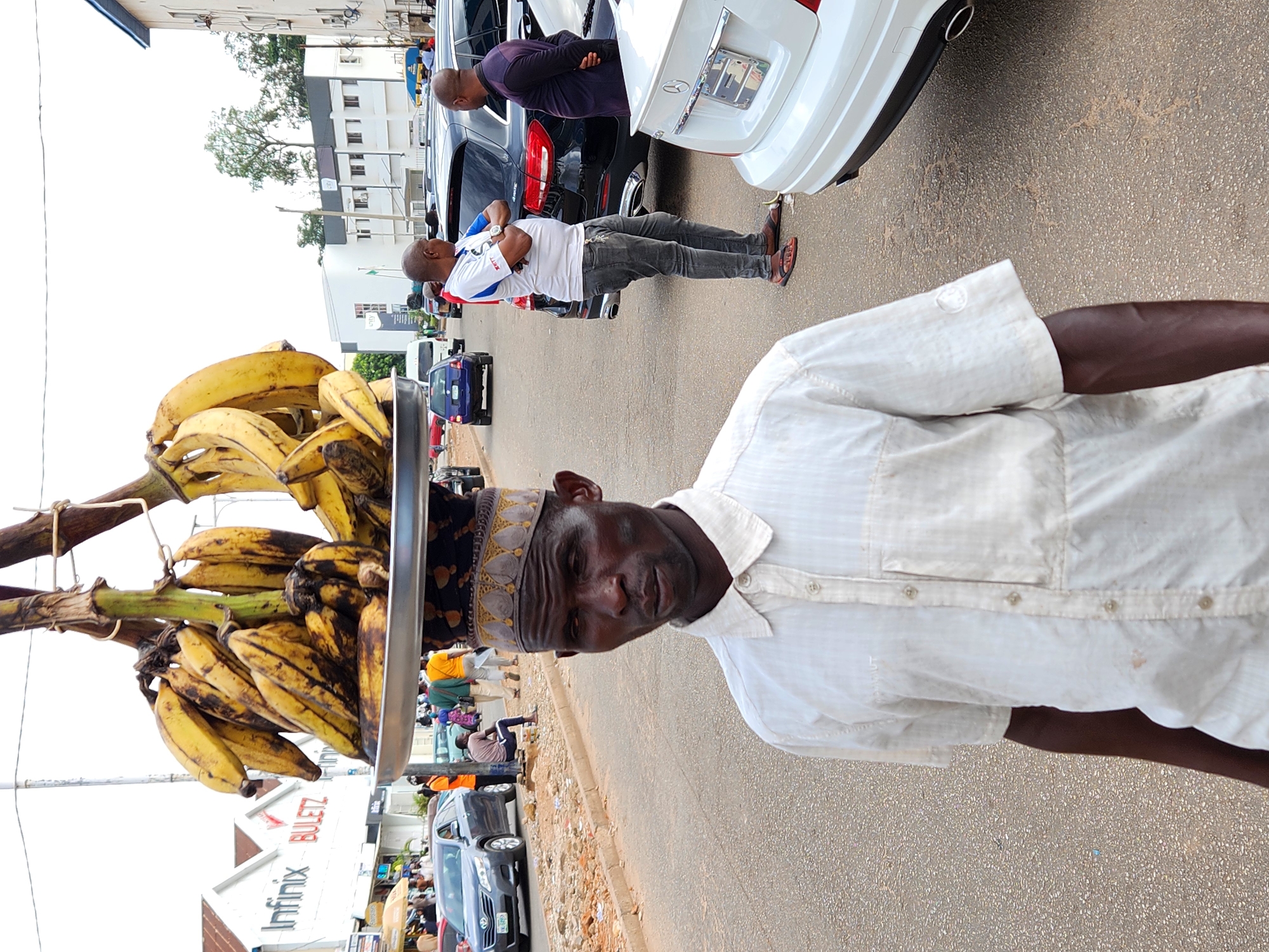 A man in a white shirt and ornamental headpiece is selling plantains on a city street in Unknown, Unknown. He carries a tray on his head piled high with ripe plantains. Behind him, people stand by parked cars on the side of the road. There is a building with an "Infinix BULETZ" sign and other businesses visible in the background.