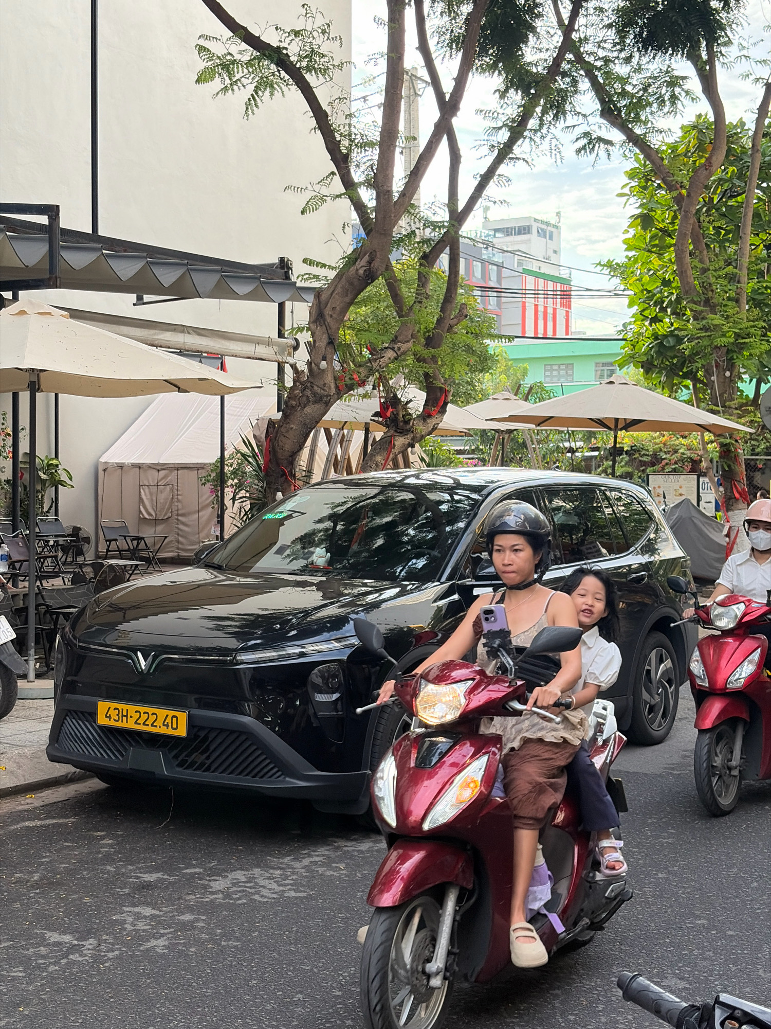 In a street scene in Da Nang, Vietnam, a woman wearing a black helmet is riding a red scooter with a young girl seated behind her. The woman is looking forward, and the girl is smiling and looking back. They are both dressed in casual clothing. Behind them, a black SUV with a Vietnamese license plate "43H-222.40" is parked on the side of the road. The street is paved, and there are trees and buildings visible in the background, suggesting an urban environment. The lighting indicates it is daytime.