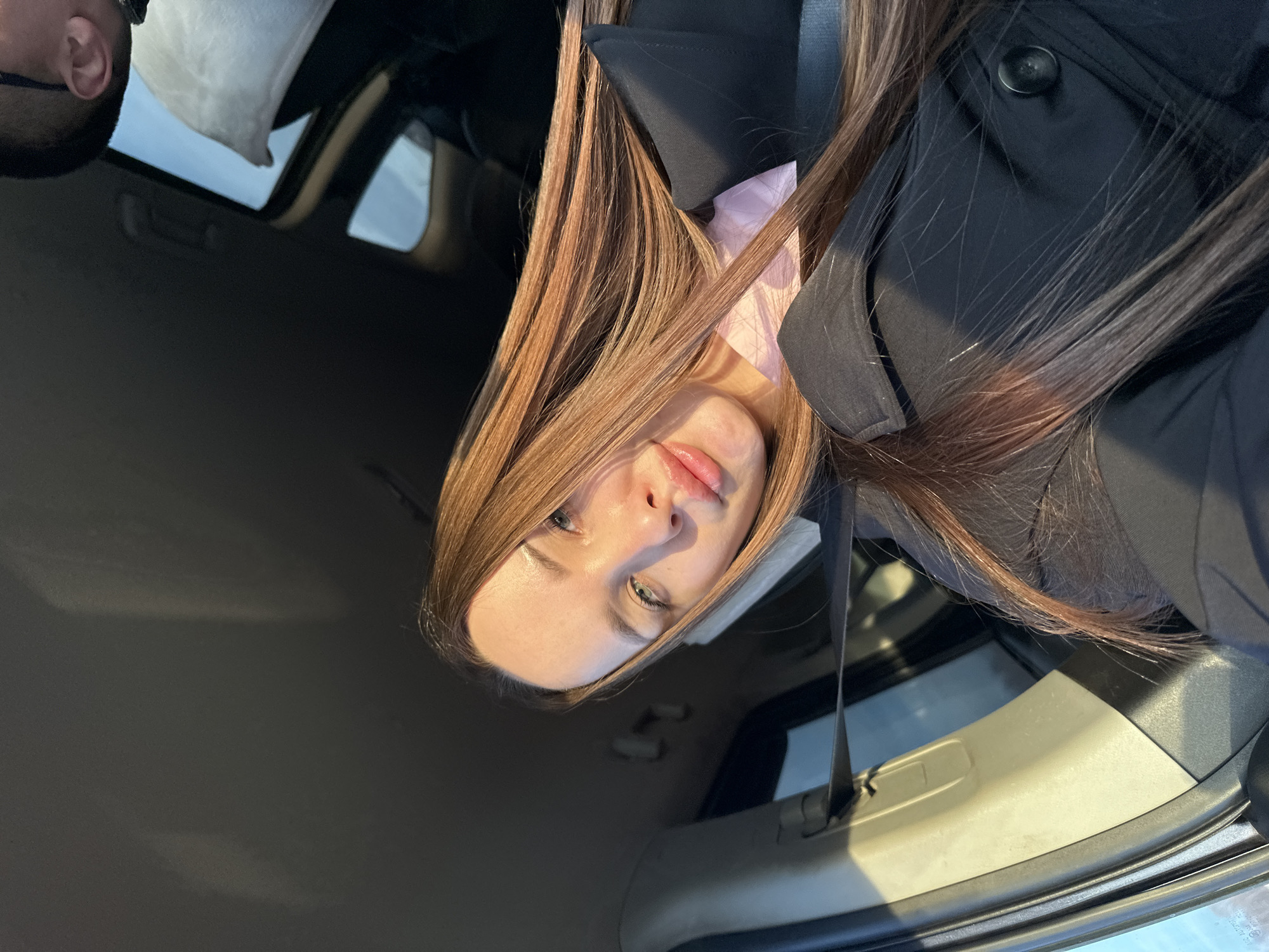 The image displays the interior of a vehicle, primarily featuring a female subject capturing a selfie. The female has long, straight, light brown hair flowing over her shoulders and chest. She possesses light-colored eyes and full lips. Her attire consists of a black collared garment, potentially a jacket or coat, layered over a visible light pink top. A black seatbelt is positioned diagonally across her torso. Strong natural sunlight illuminates the right side of her face and hair. In the upper right quadrant of the image, the top of a male individual's head, including his right ear and dark-framed glasses, is partially visible, indicating his presence in the rear of the vehicle. He appears to be resting against a white, possibly cushioned, object, wearing a dark shirt. Above the male subject, a vehicle sunroof is open, revealing a clear blue sky. The car's interior features a dark overhead lining and a light-colored B-pillar. The scene is located in Pirogovo, Russia.
