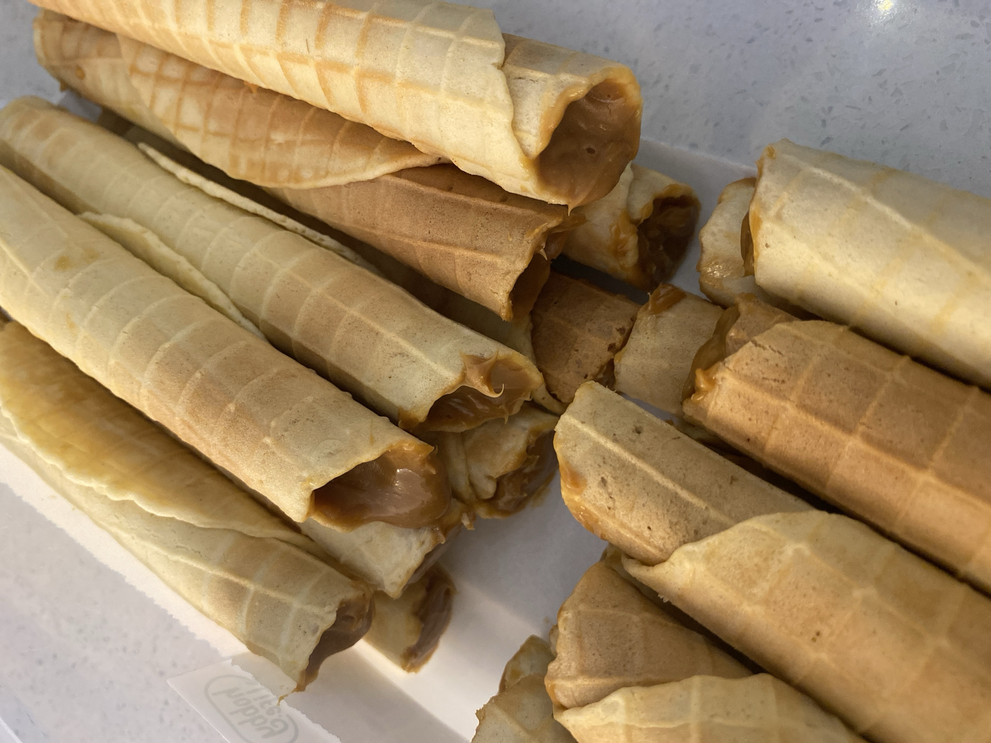 The image provides a close-up view of numerous golden-brown, tubular pastries, likely waffle rolls, arranged within a white container or on a light-colored surface. Each pastry features a distinctive waffle-like grid pattern on its exterior and is filled with a creamy, light brown substance, resembling dulce de leche or caramel. This sweet filling is visible at the open ends of many of the rolls, and in some instances, appears to be slightly oozing out.
The pastries vary slightly in hue, with some appearing a lighter golden-beige and others a deeper, richer brown, suggesting subtle differences in their preparation or baking time. They are piled casually, creating a sense of abundance and appeal. The background consists primarily of the white container, with a subtle textured, light grey countertop visible in the upper right.
There are no people present, and no specific activity is captured other than the static display of the food, implying they are ready for consumption or serving. The lighting suggests an indoor setting, likely under artificial light. No explicit location cues specific to Novi Sad, Serbia, are visible within the frame, but the pastries themselves are a type of sweet treat found in many cultures, including Eastern European regions. A partially visible white label at the bottom left bears stylized text, which appears to read "Moddor" or "Moddori", possibly indicating a brand or manufacturer on the packaging.