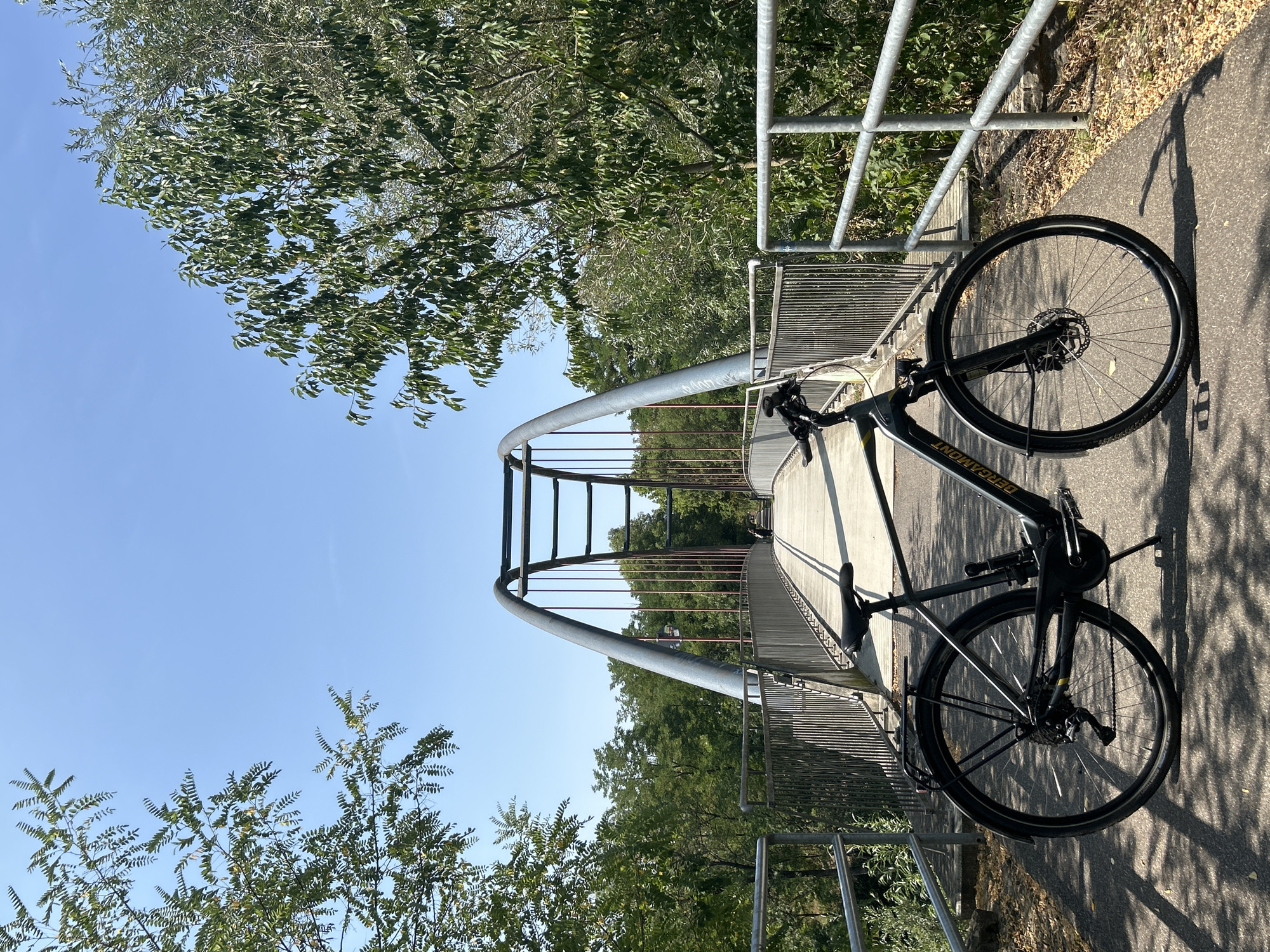 An electric bicycle, a Bergamont E-Ville model, is parked on a paved pathway on a pedestrian bridge in Oedheim, Germany. The bridge has a distinctive arch structure with metal cables supporting the walkway, which is bordered by a metal railing. Trees and foliage line the sides of the pathway leading up to and beyond the bridge. The sky is clear and blue, indicating a sunny day. In the background, a person is visible walking further down the bridge. The bicycle is positioned in the foreground, facing slightly to the left, with its kickstand deployed. Shadows of the trees and the bridge's railing are cast across the pathway.