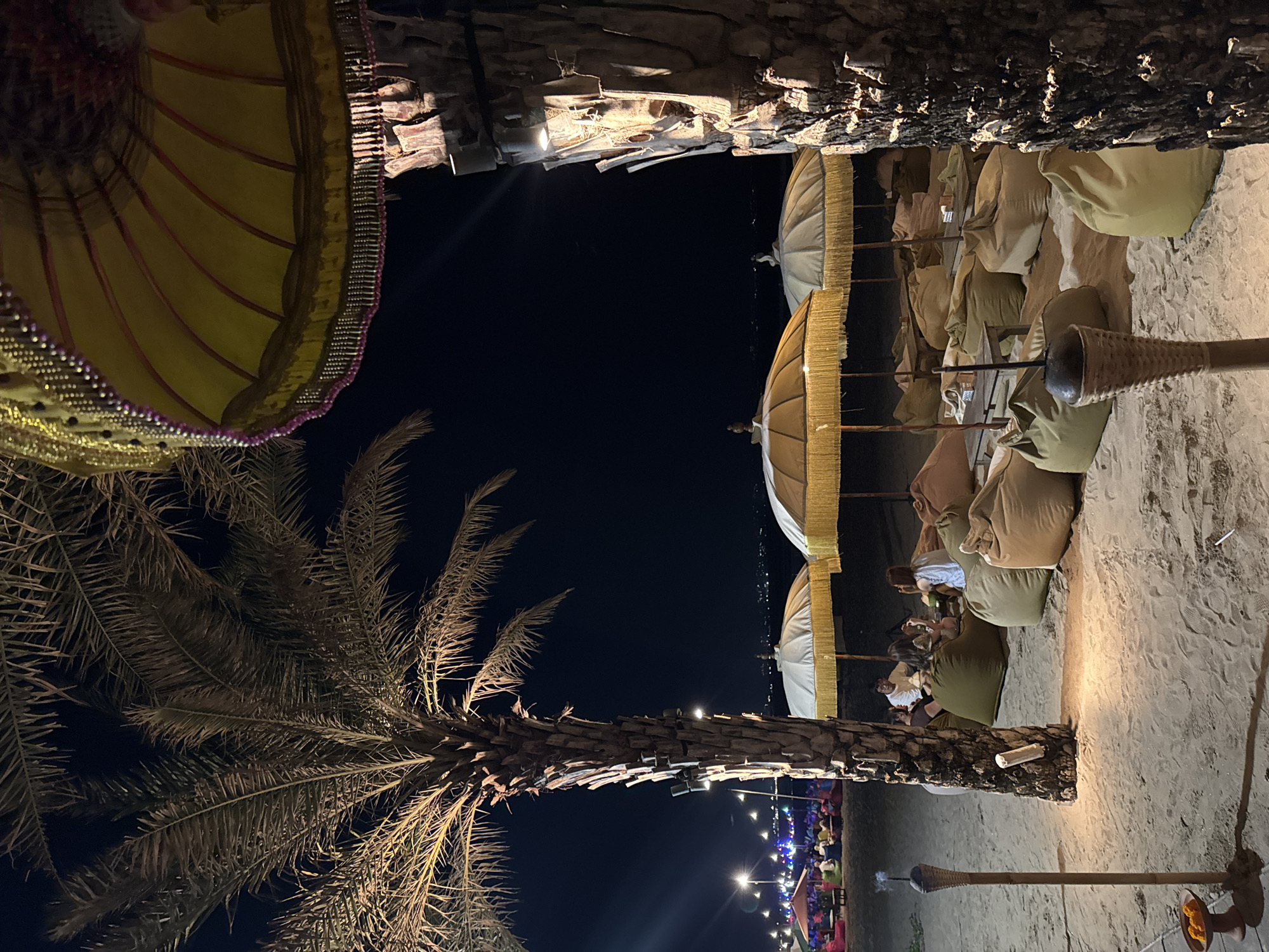 This is a low-angle, medium-distance, eye-level, night-time, outdoor shot of a beachside seating area in Kuta, Indonesia. The foreground features the textured bark of a palm tree on the right and a torch with a small offering dish in the lower left corner, casting a warm glow on the sandy ground. The middle ground displays a relaxed seating arrangement with numerous large, light-colored beanbag chairs scattered on the sand, some with small wooden tables. Several beach umbrellas with yellow fringed edges provide shade over the seating areas. A group of four individuals, two men and two women, are seated in the beanbags under one of the umbrellas, interacting with each other. In the background, more beach umbrellas and seating areas are visible, along with lights strung across the beach, suggesting a vibrant evening atmosphere. The distant horizon shows the dark outline of the sea and possibly buildings or distant lights on the shore. The overall scene depicts a casual, illuminated beach venue at night.
