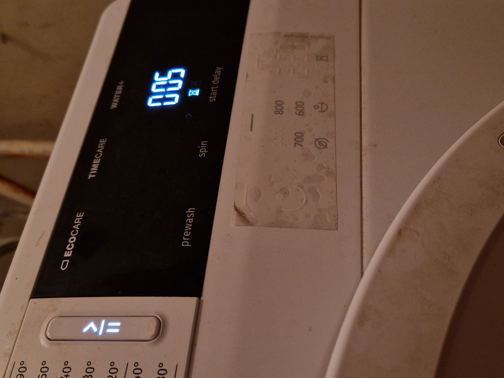 A close-up, eye-level, indoor, medium shot captures a section of a white washing machine's control panel. The panel is predominantly black with white text and illuminated blue digital displays. On the left, a vertical temperature dial shows degrees Celsius: 90°, 60°, 40°, 30°, 20°, 40°, 30°. Next to the dial is an illuminated white button with a blue symbol that resembles an upward arrow over an equals sign. The black section of the panel features text labels: "ECOCARE", "TIMECARE", and "WATER+". Below "TIMECARE", the text "prewash" is visible, and below that, "spin" and "start delay". A blue digital display shows "005", with a small hourglass icon beneath it. To the right of the digital display is a faded label with several icons and numbers, including "1000", "800", "700", "600", and symbols that appear to represent no spin, a droplet, and a timer. The washing machine's surface is visibly dirty with smudges and dust. The background is blurred and appears to be an interior space with some pipes or wires visible. The image does not contain any contextual information regarding the city of Raduvene, Bulgaria.