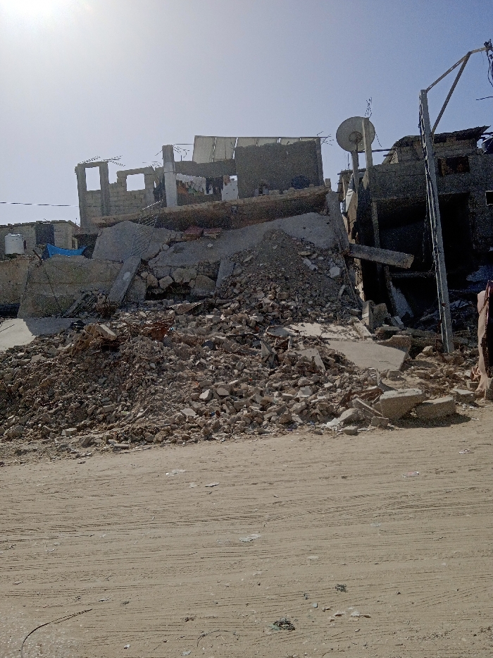The image depicts a scene of extensive structural damage in Khan Yunis, Palestinian Territory, under a bright, clear sky. In the foreground, an unpaved, dusty ground with tire tracks leads to a large, prominent mound of concrete rubble, fragmented bricks, and twisted rebar, originating from collapsed buildings. Behind this debris, multiple residential structures are visible in various states of disrepair. To the left, a multi-story building shows significant damage, with its upper floors partially destroyed and exposed, revealing internal walls and openings. Laundry hangs on a line between supporting structures on one of these damaged upper floors. A blue tarp and a white water tank are visible near the base of structures further left. To the right of the central rubble pile, another building appears heavily damaged, with large sections of its facade missing. A satellite dish is mounted on a structure near the center-right. A tall metal utility pole with attached wires stands upright on the far right. No people or active interactions are present in the static scene.