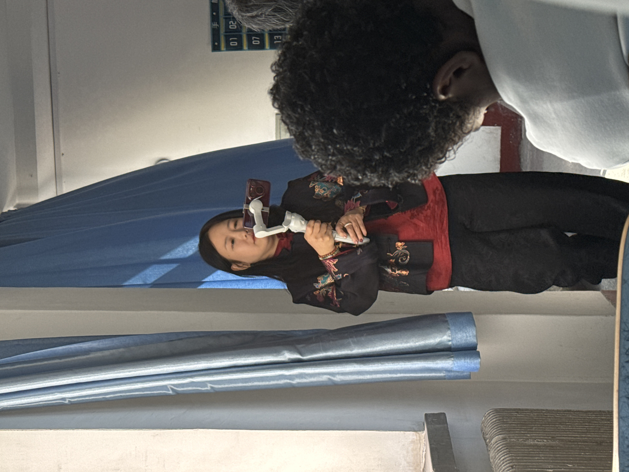 This indoor image captures two individuals, with one prominently in focus. In the center, an East Asian woman is holding a smartphone attached to a white gimbal or stabilizer, seemingly operating it for recording or livestreaming. She is dressed in traditional attire, featuring a black jacket adorned with colorful embroidery over a red garment, paired with black trousers. Her gaze is directed slightly upwards and to the left, with a neutral and focused expression.
Partially visible in the upper right is another person with dark, curly hair, seen from behind and wearing a light-colored top.
The background is dominated by blue and light blue curtains or fabric drapes. Above the woman, a light-colored wall or ceiling is visible, featuring a dark-framed board with numbers and Chinese characters. Visible text includes "手" (shǒu), "20", "01", "02", "07", and "13". A horizontal light-colored beam or shelf is also present in the mid-ground. To the lower right, a dark, textured object, possibly furniture or equipment, is partially visible.
The setting appears to be a studio or a space prepared for video content creation. The visible Chinese characters are consistent with the provided location context of Zhongtun, China. No specific time of day or weather is discernible from the indoor scene.