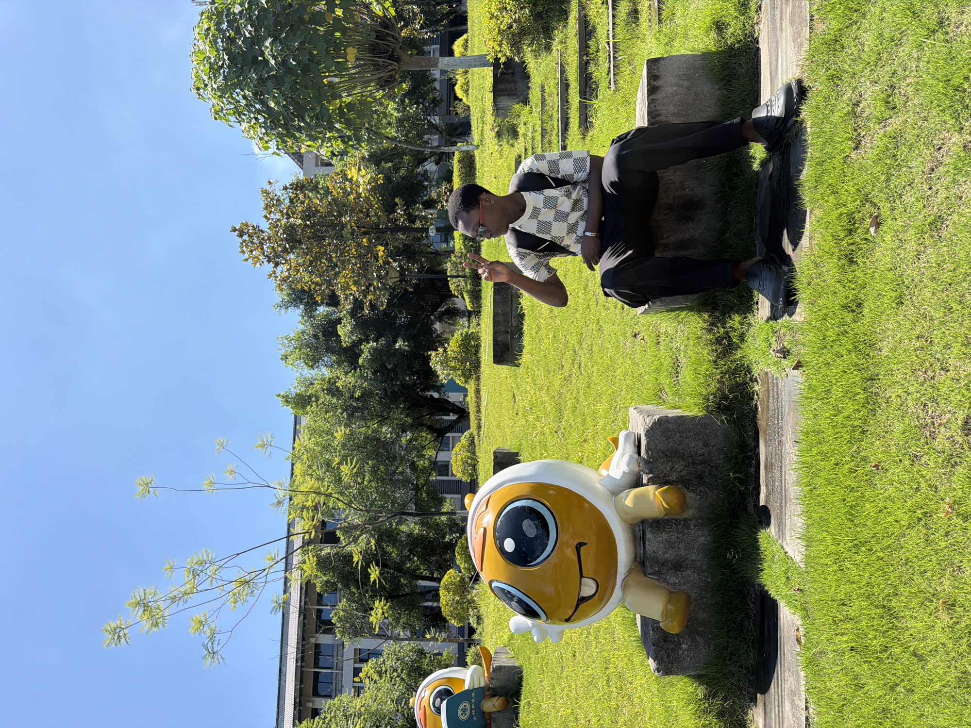 A young Black male wearing a black and white checkered shirt, black pants, and black shoes sits on a concrete block in a park. He has sunglasses on and is making a peace sign with his right hand. To his left is a large, yellow and white cartoonish statue of a character with big eyes and a wide smile. The statue is also sitting on a concrete block. The park has green grass, trees, and a building in the background. The image appears to have been taken on a sunny day. The background building and signage on the statue suggest the location is the Jiangjin District of Chongqing, China, likely within a university or public park, possibly affiliated with the Chongqing University of Science and Technology given the insignia on the book held by another statue to the left, which is partially visible.