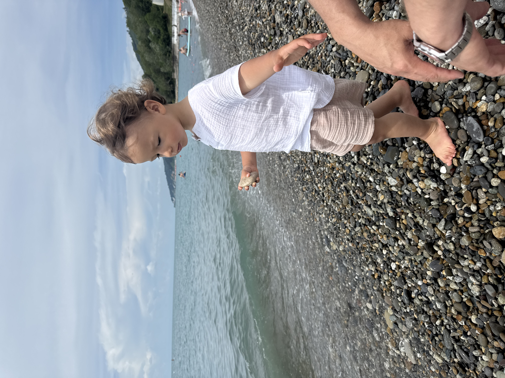 A young child with curly brown hair stands on a pebble beach, looking down at a small stone in their hand. The child is wearing a white, textured short-sleeved shirt and light pink shorts. To the right, a person's arm with a watch on their wrist is visible. The sea is to the left, with gentle waves lapping the shore. In the background, there are people swimming and on paddleboards in the water. A forested hillside is visible beyond the beach. The sky is overcast with some clouds. The location is identified as a beach in Sochi, Russia.