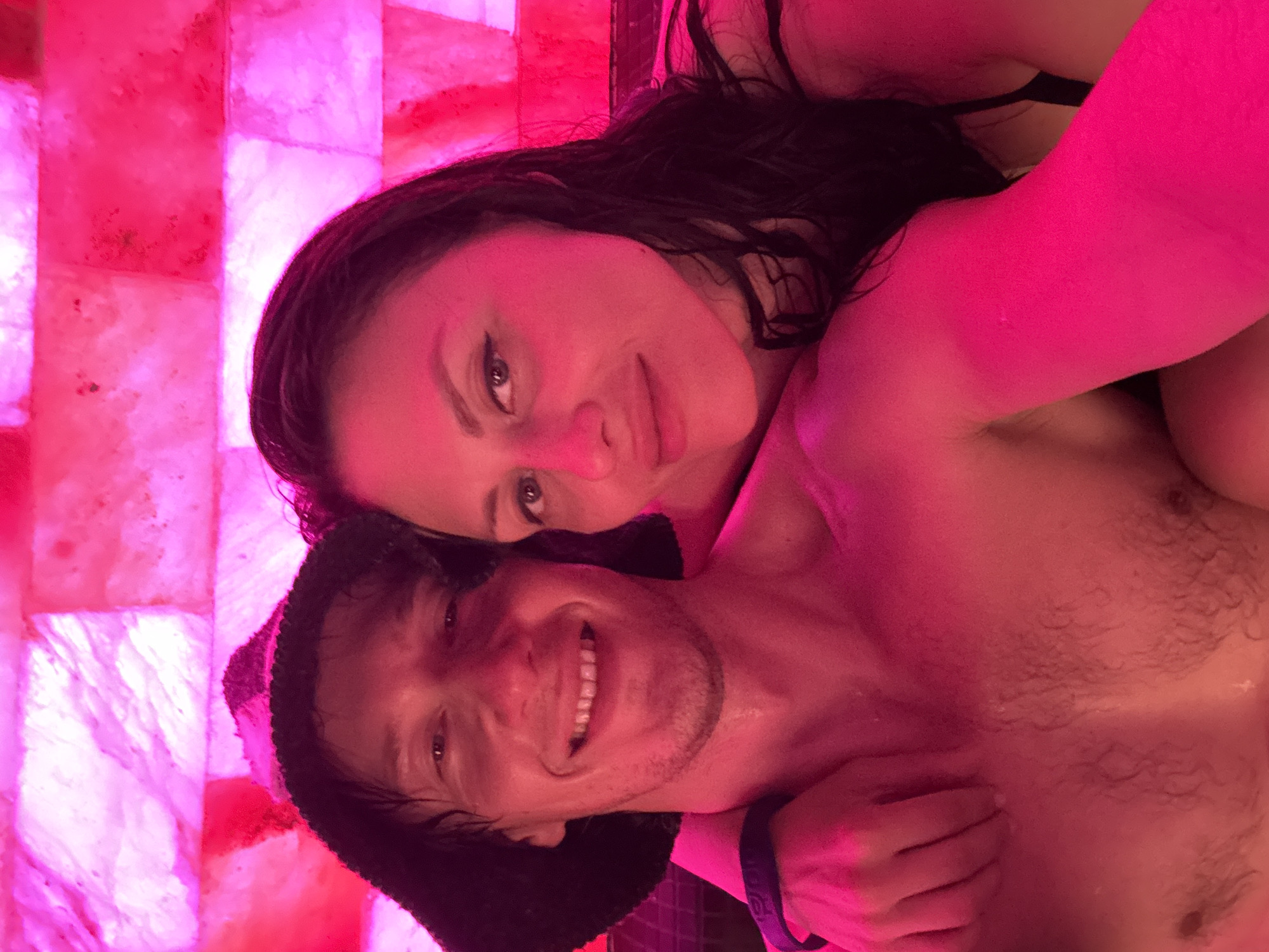 A close-up, angled portrait features a man and a woman embracing in a brightly lit, pink-hued setting. The background is a wall composed of translucent, rectangular salt blocks, internally illuminated with a vibrant pinkish-red light, strongly suggesting a salt room, sauna, or spa environment.
The woman is positioned above the man, looking directly at the camera with a calm expression. She has long, dark, possibly damp hair, and dark eyes with visible eyeliner. She appears to be wearing a dark top, likely a bra or swimsuit, and her skin is illuminated by the pink light.
The man is below the woman, also looking at the camera, with a wide, joyful smile revealing his teeth. He has short dark hair, some stubble on his chin, and is shirtless, showing his chest. His hair and skin also appear damp or glistening, consistent with a warm or humid environment. He wears a dark, possibly fuzzy, beanie or headwear and a dark wristband on his right wrist. His arm is around the woman's shoulder or back.
Both individuals convey happiness and intimacy. The predominant pink lighting creates a warm, atmospheric, and somewhat romantic ambiance around them. No visible text is discernable on the wristband or in the background.