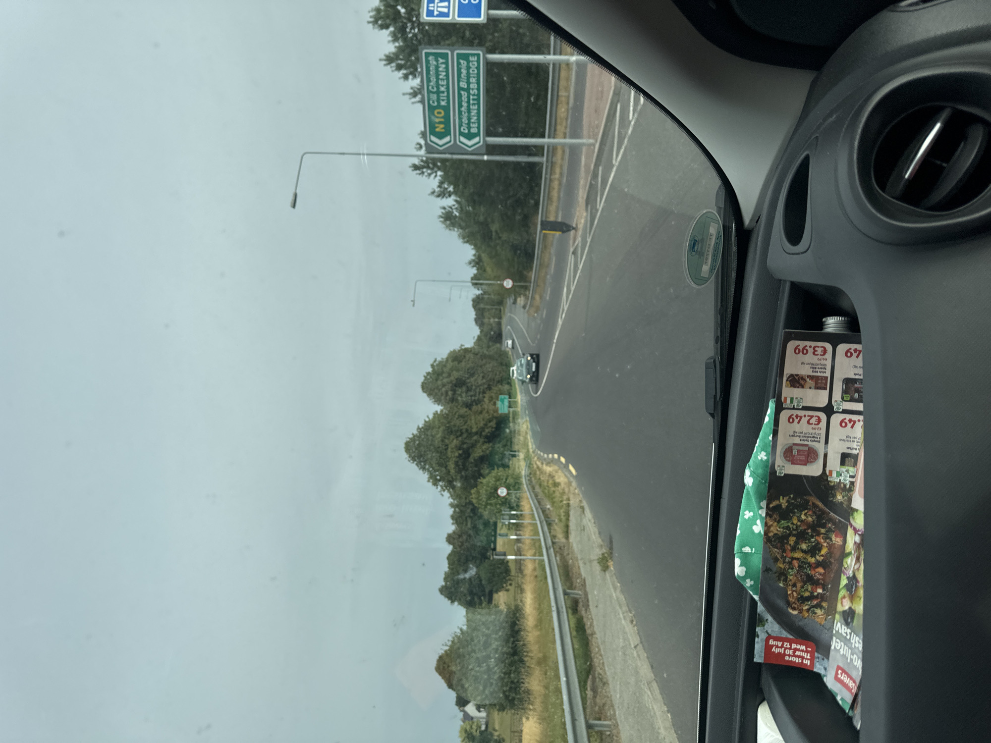 The image is a first-person view from inside a car, looking forward through the windshield. The car is on a road, with trees and fields on either side. There are road signs indicating directions to "N10 Cill Chainnigh KILKENNY" and "Draíocht Bhíde BENNETTSBRIDGE". The road appears to be in Ireland. In the foreground, on the dashboard, are some flyers and a green patterned cloth. The sky is overcast.