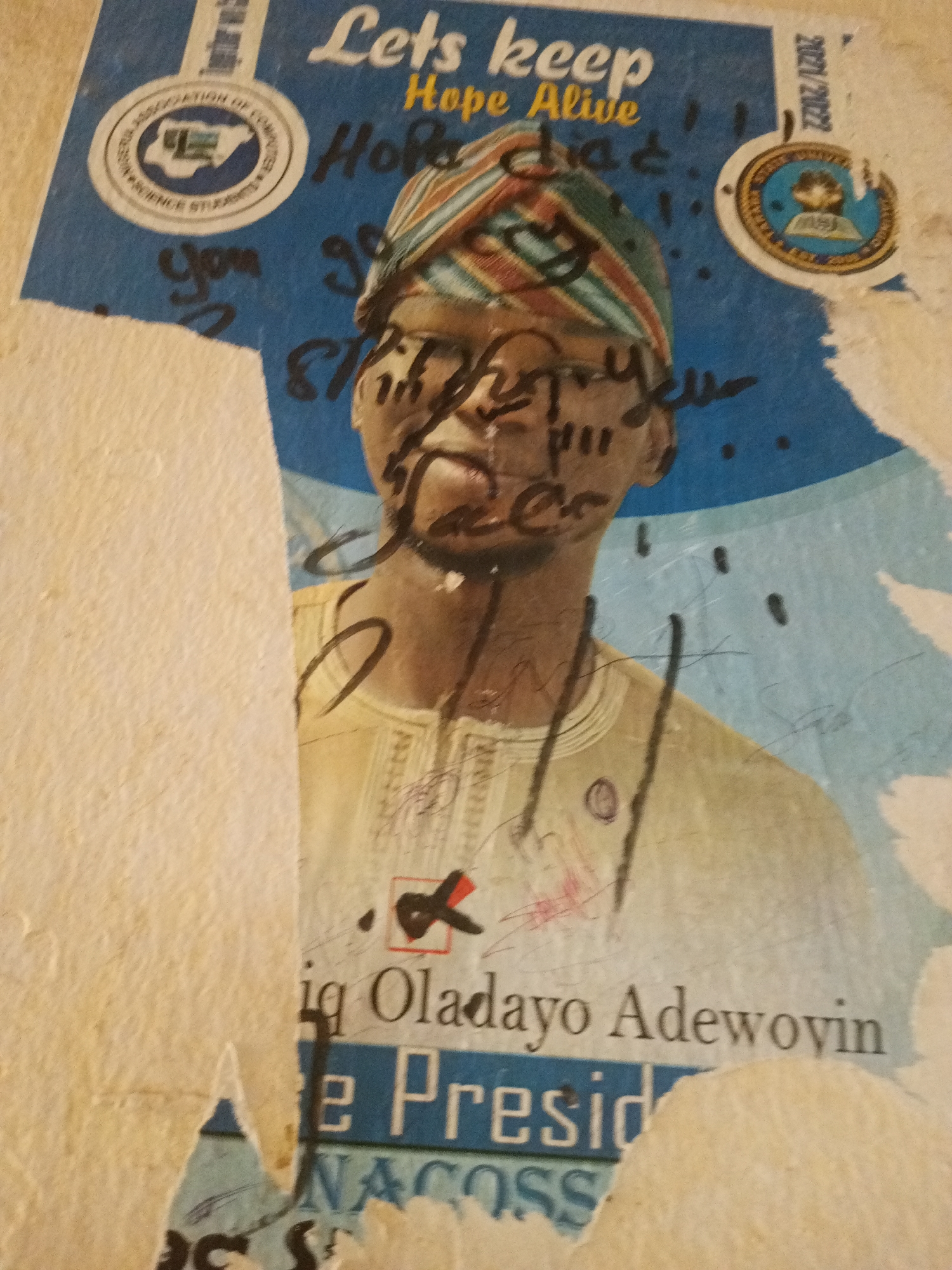 The image is a close-up, slightly blurry, vertically oriented photograph of a torn poster on a textured, light-colored wall, likely located in Jalingo, Nigeria. The poster features a blue background with a portrait of a young man wearing a colorful striped headwrap and a light-colored, embroidered shirt. He has dark skin and is looking slightly upwards. Black marker scribbles obscure parts of his face and other areas of the poster. Text on the poster is partially visible, including "Lets keep Hope Alive" and "2021/2022". Below the man's portrait, some text is legible, reading "Oladayo Adewoyin" and "e Presid" and "NACOSS". There are also circular logos with text and images on the poster, one on the left with "ASSOCIATION OF COMPU..." and another on the right with a person inside a circle. The poster is damaged with rips and peeling paper, revealing the wall beneath. The overall impression is one of a defaced election or campaign poster.