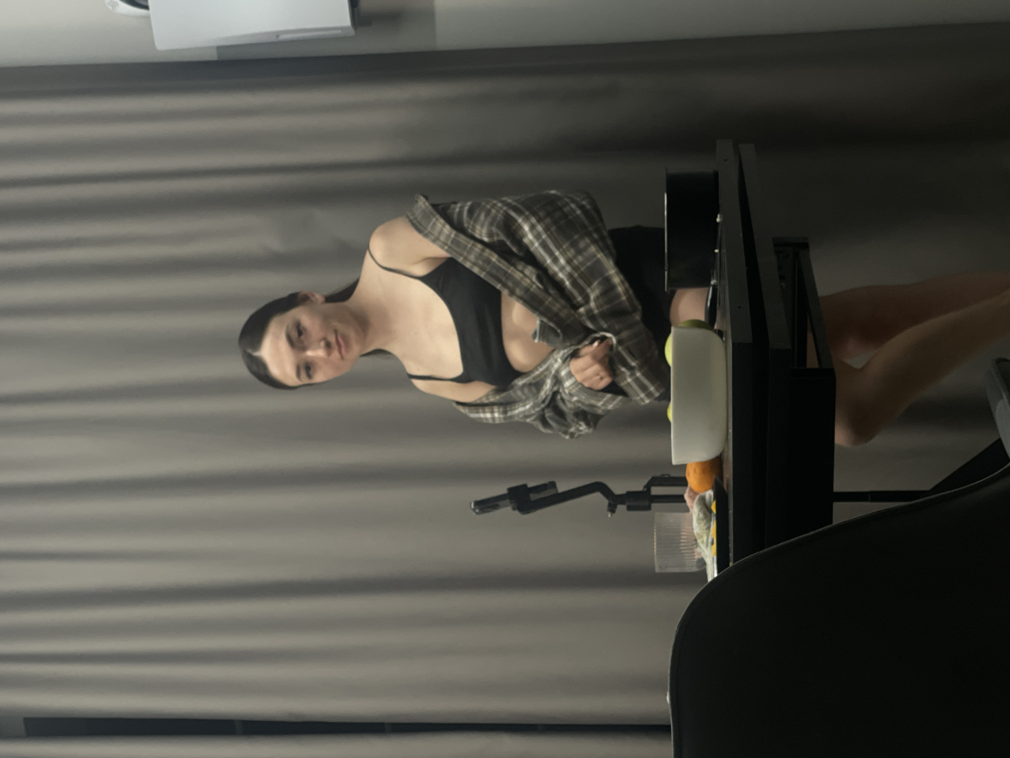 A young woman in a black top and flannel shirt sits to the right, partially obscured by a black desk. The desk has a white bottle and other items on it. A black adjustable microphone stand is visible. A black office chair is in the foreground. The background is a light grey curtain. The image does not provide sufficient information to determine if the location is in the city of Эстачи, Russia.