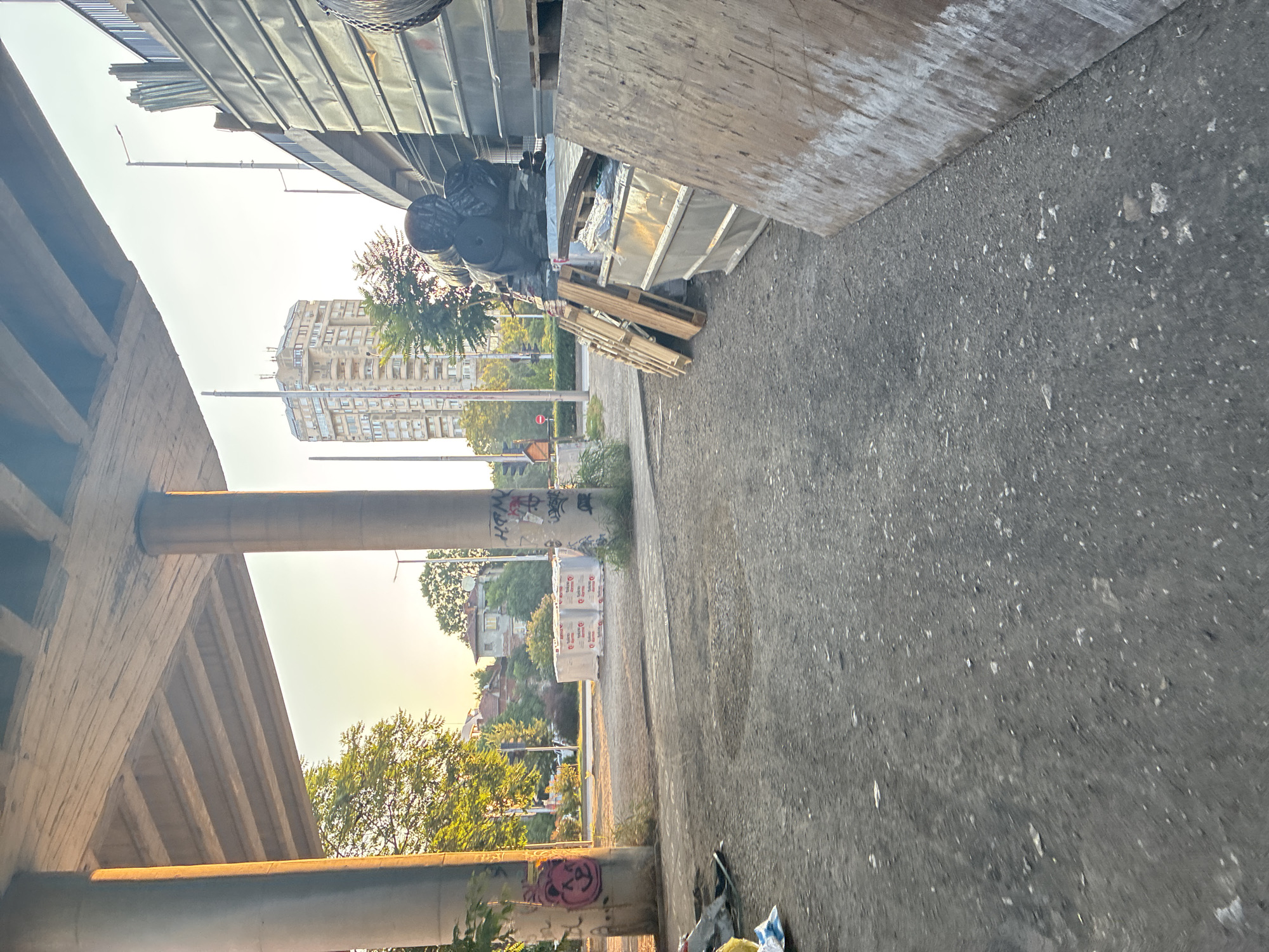 The image depicts an underpass in Plovdiv, Bulgaria, likely near a construction or industrial zone. The scene is characterized by a rough asphalt ground, two large concrete pillars supporting an elevated structure, and various construction materials. Stacked white packages, possibly of insulation or building materials, are visible in the mid-ground. To the right, a large metal structure, possibly part of an ongoing construction project, is present, along with wooden pallets and large rolled-up materials covered in black plastic. A tall, modern building stands in the background, suggesting an urban environment. The lighting indicates it is either early morning or late afternoon, with sunlight casting long shadows and highlighting the textures of the concrete and ground. Graffiti is present on the concrete pillars. The overall impression is one of an unfinished or transitional urban space.