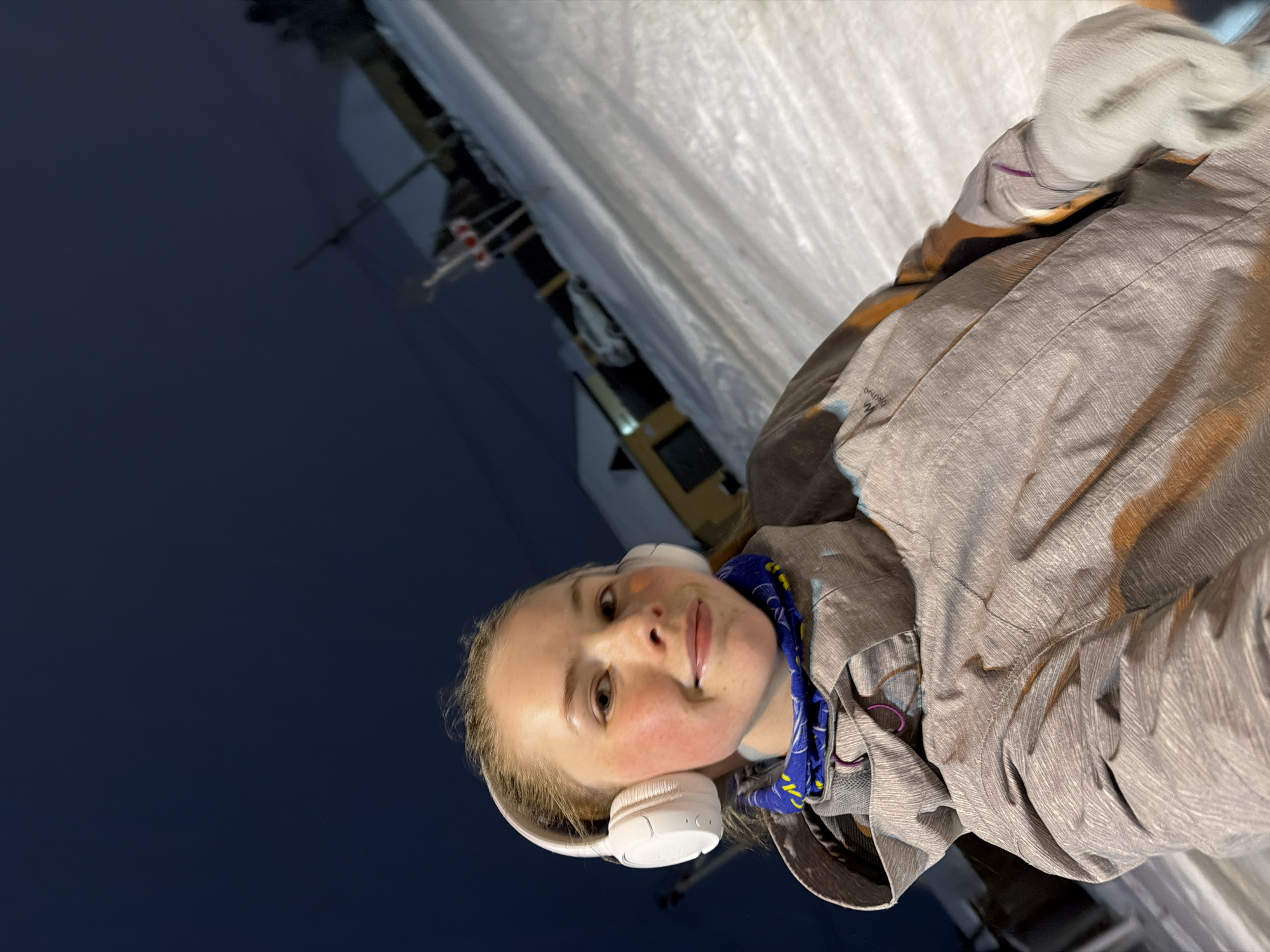 A young woman is centered in the frame, wearing white headphones and a gray jacket. She is looking at the camera with a slight smile. Her cheeks are flushed. A blue patterned scarf is visible around her neck. The background is divided between a dark navy blue sky and a snow-covered surface. A structure with flags and a ski lift is visible in the distance, suggesting a ski resort setting. The image appears to be taken at night or in low light conditions. Without additional information, it is not possible to confirm the specific location within Berezniki, Russia.