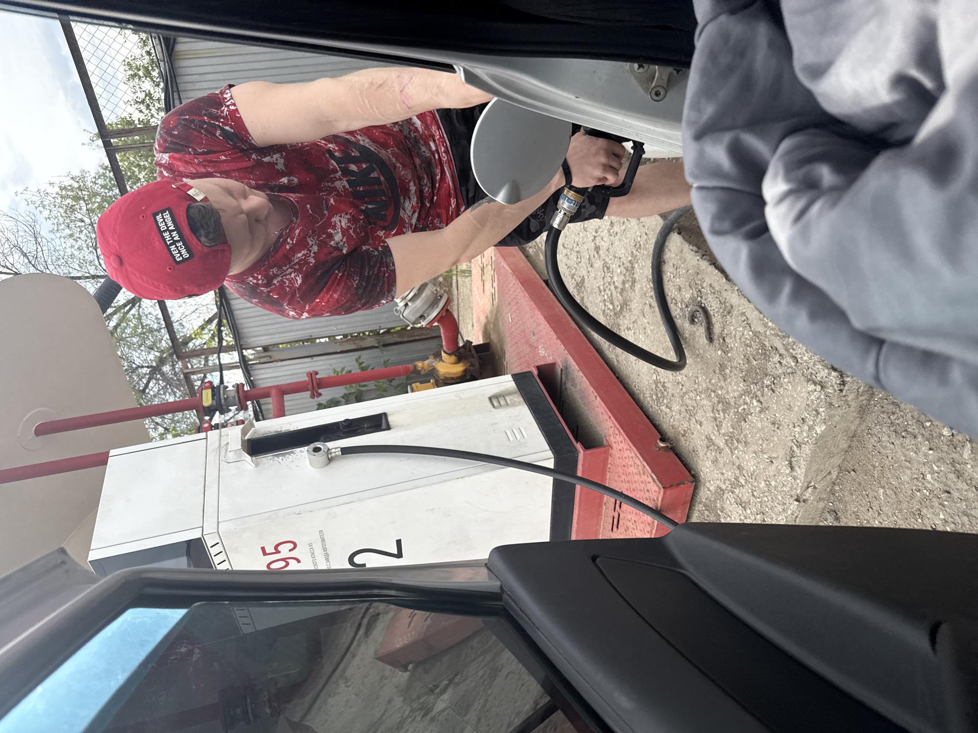 A young person wearing a red baseball cap and a red and white tie-dye t-shirt with a Nike logo is fueling a silver car at a gas station in Samara, Russia. The gas pump is white with red lettering indicating "95" and the number "2". A black hose extends from the pump to the car's gas tank, which is open. The ground is paved with concrete and small stones. In the background, a metal fence and trees are visible. The scene is captured from inside another vehicle, showing part of its interior in the foreground.