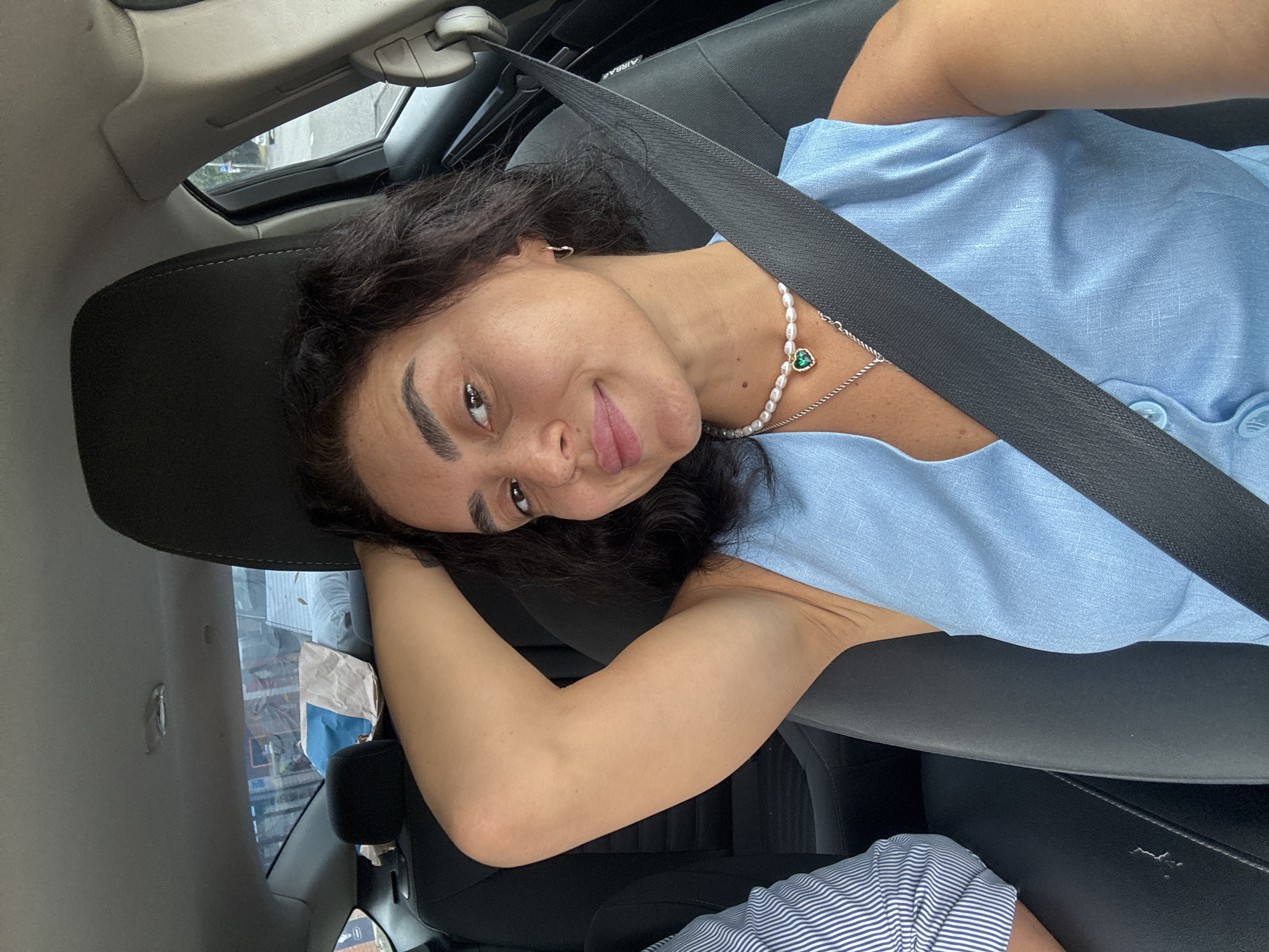 A medium shot shows a woman with dark, curly hair and fair skin from a first-person perspective, sitting in the passenger seat of a car. She is looking at the camera with a slight smile and has her right arm raised behind her head. She is wearing a light blue sleeveless dress with visible buttons and a pearl necklace with a green heart-shaped pendant. A black seatbelt crosses over her chest. To her left, partially visible, is a person wearing a blue and white striped shirt. The background shows the interior of the car, including the headrest and part of the dashboard, as well as a window revealing a blurred outdoor scene that appears to be a street in Tbilisi, Georgia, with buildings and some foliage visible. The lighting suggests it is daytime.