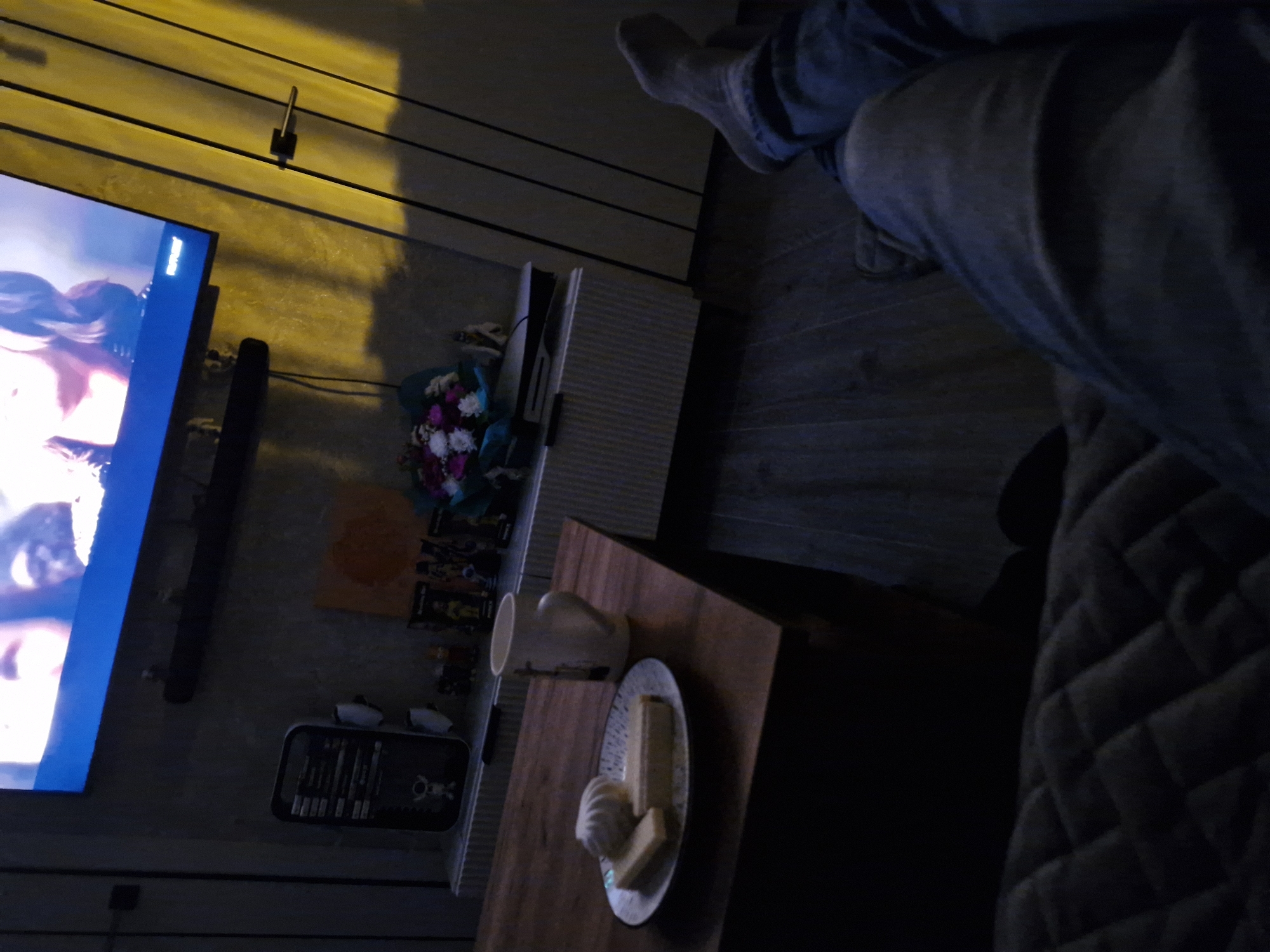 A low-angle, medium shot shows a person's legs and feet wearing gray socks and blue jeans, resting on a quilted surface. The background is dimly lit, with a television screen displaying an image, a wooden table with a plate of pastries and a mug, and decorative items on a shelf. The room has a textured wall and a wooden floor. The scene appears to be an indoor living space.