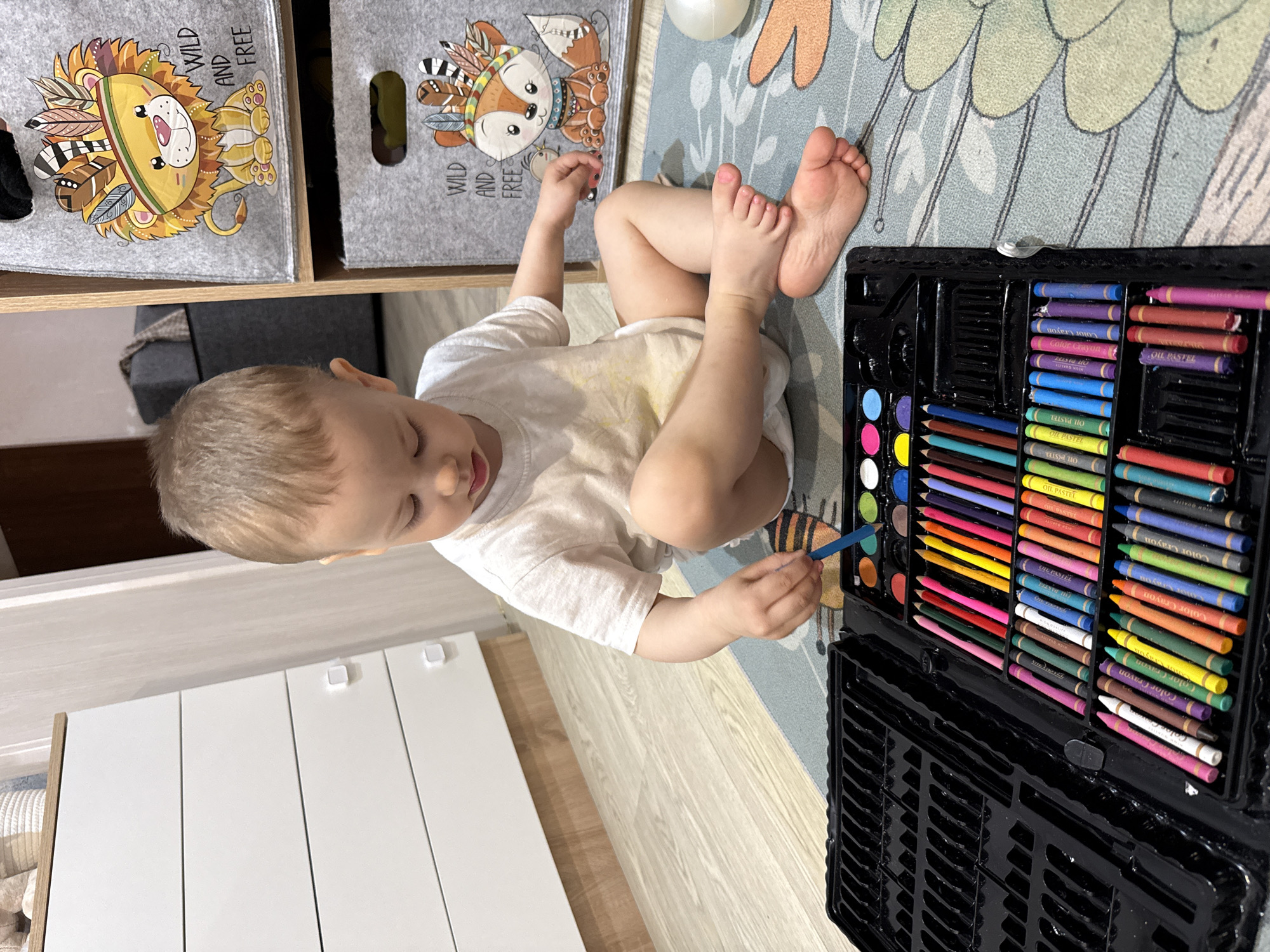 A child sits cross-legged on a patterned rug in an indoor setting, possibly in Rusanovo, Russia. The child wears a light-colored shirt and appears to be using a paintbrush with a black palette containing watercolors and oil pastels. The environment includes white furniture with simple square handles, and storage bins decorated with cartoon animals (lion and fox) and the words "WILD and FREE."
