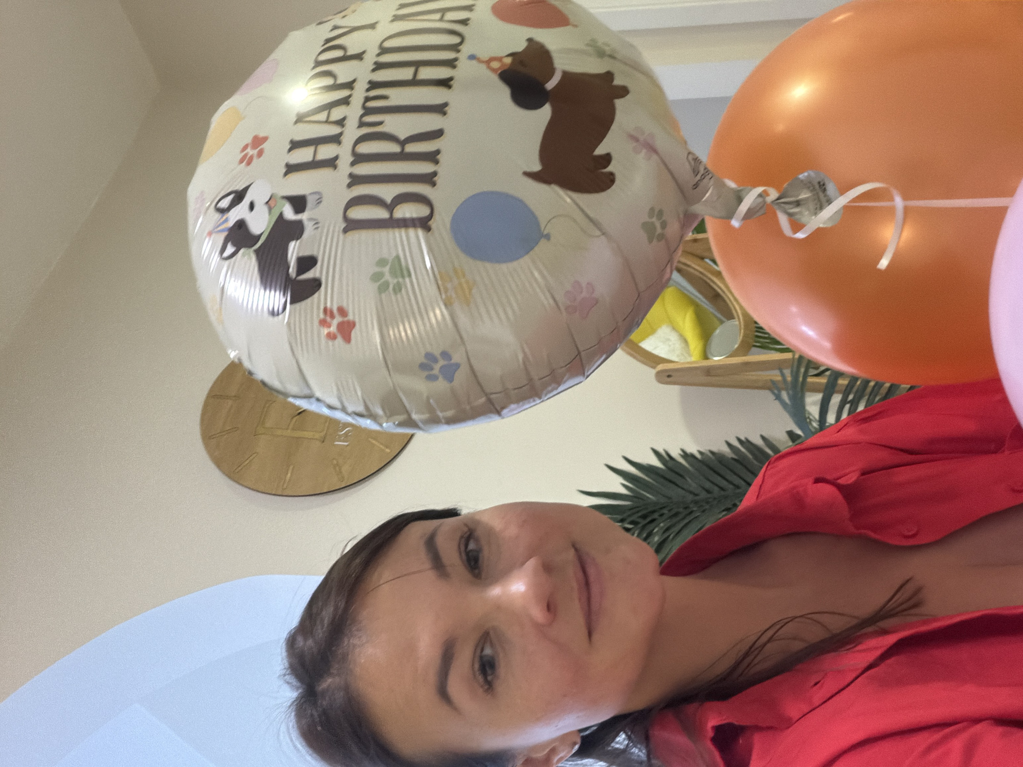 A woman is taking a selfie in a room, possibly a living room, with birthday decorations. She is wearing a red shirt and has her hair pulled back. She is smiling at the camera. In the background, there are balloons, including one that says "Happy Birthday" with images of dogs. There is also a clock on the wall and a plant. The setting appears to be a home environment. There is no visible indication of the location being in Budva, Montenegro.