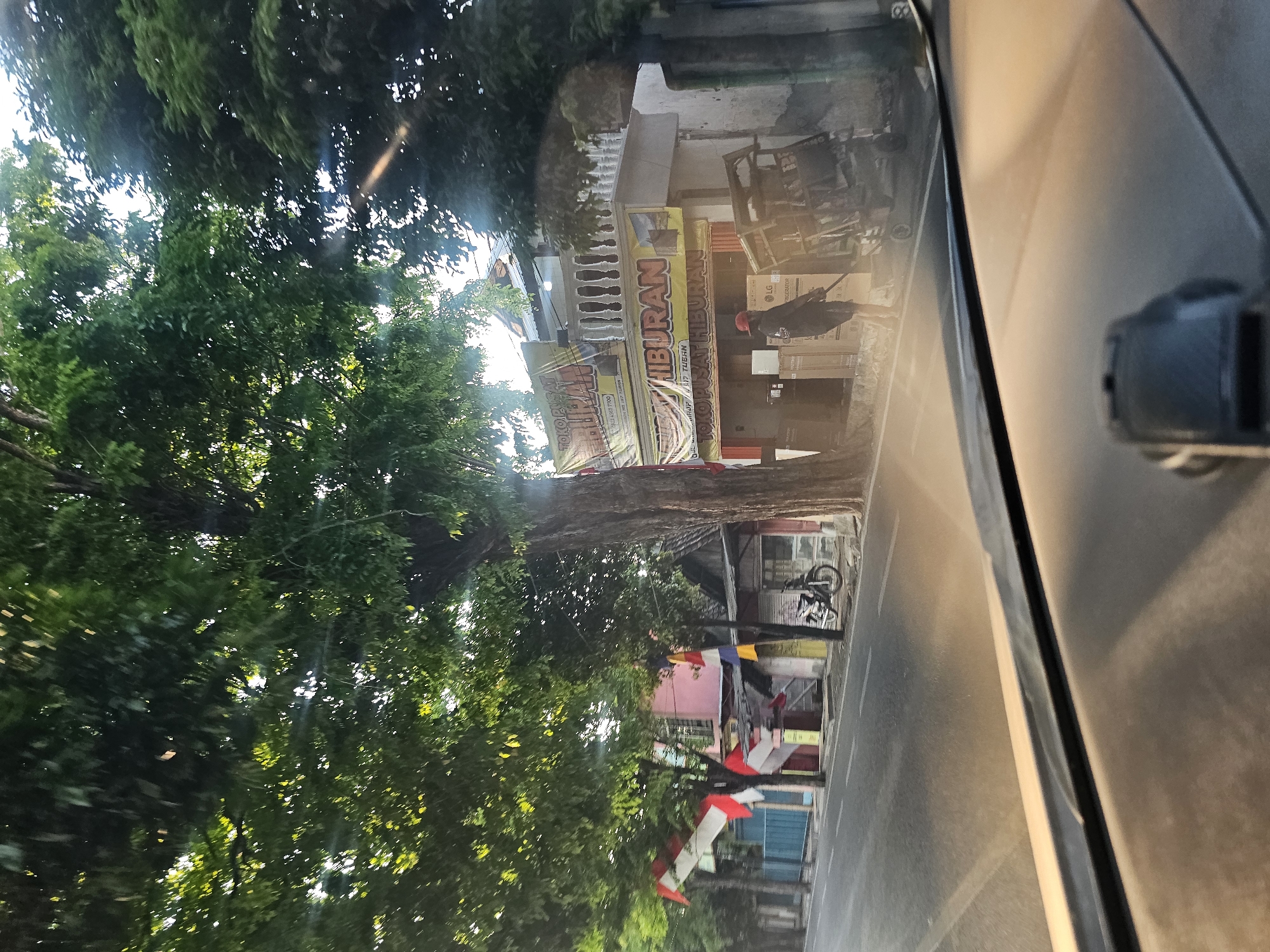 The image depicts a street scene in Tuban, Indonesia, likely in the early morning or late afternoon due to the soft lighting. Tall trees with dense green foliage overhang the street, casting dappled sunlight. In the background, a two-story building with a white railing on the upper level is visible. A prominent banner advertising "TOKO PUSAT HIBURAN" (Entertainment Center Shop) in yellow and red is displayed across the front of the building. Below the banner, several large boxes, possibly appliances, are stacked outside. A person wearing a red cap and dark clothing stands near the boxes, holding what appears to be a broom or a long stick. To the right of the person, a metal cart with wheels is positioned. In front of a building to the left, a motorcycle is parked. Indonesian flags, predominantly red and white, are displayed on poles along the street. The foreground shows the interior of a vehicle, with the dashboard and windshield visible. The street itself is paved and marked with white lines. The overall impression is of a commercial street in a tropical urban environment.