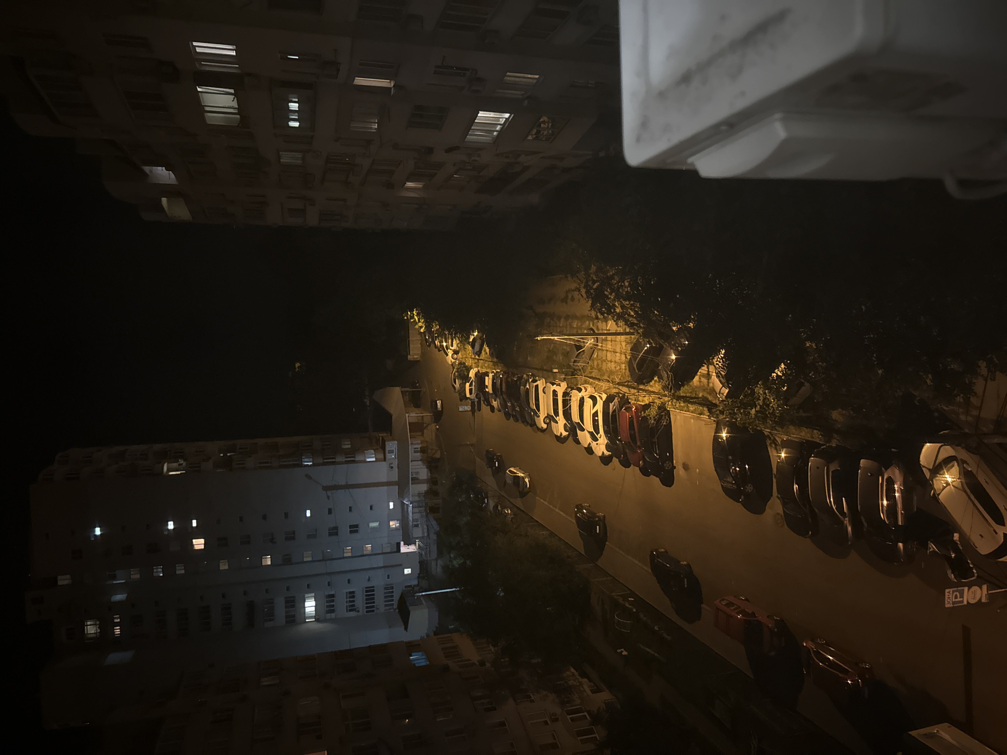 The image depicts a nighttime urban scene, likely in an apartment complex in Unknown, Unknown. Two tall apartment buildings flank a street lined with parked cars. The buildings are illuminated by internal lights, with some windows glowing warmly and others dark. The street is dimly lit, casting long shadows from the cars and trees. A row of cars is parked along the right side of the street, and several more are parked on the left. The overall atmosphere is quiet and still, typical of a residential area at night. The foreground shows a white object, possibly an air conditioning unit or part of a balcony, in sharp focus.