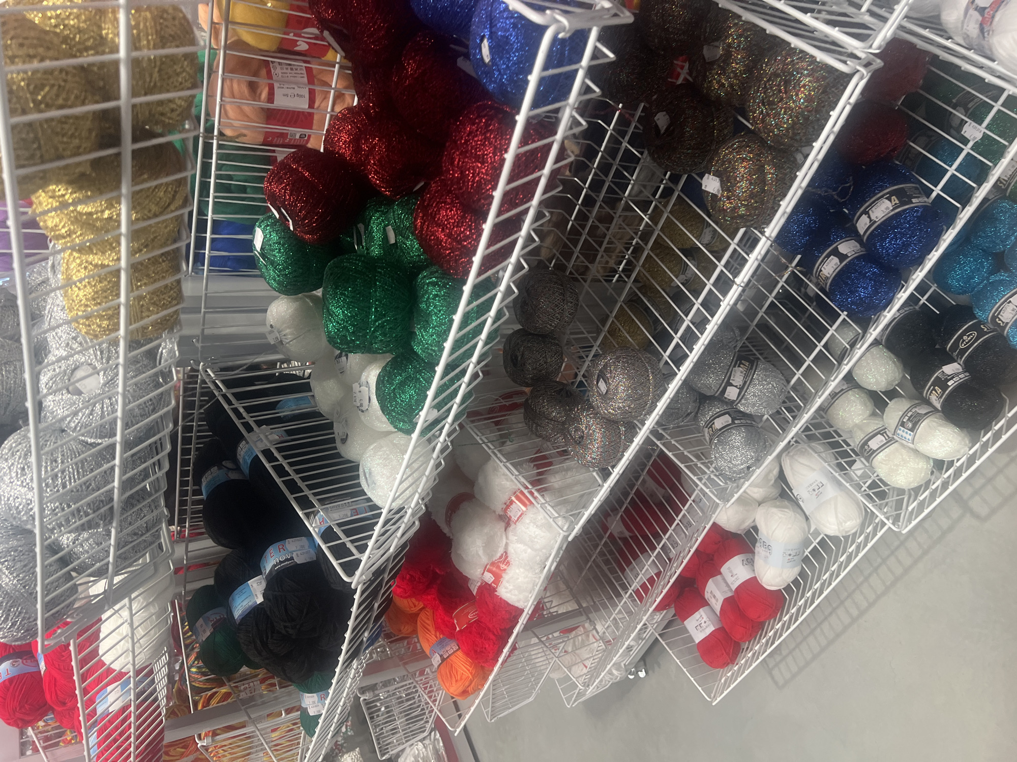 The image depicts a store display of colorful yarn skeins, likely taken in a retail setting in Burgas, Bulgaria, based on the brand names visible on the labels. The yarn is organized in multiple rows on a white metal shelf rack. The yarn skeins are assorted in colors like gold, silver, red, green, white, black, blue, and various shades of brown and gray. The yarn appears to be sparkly.