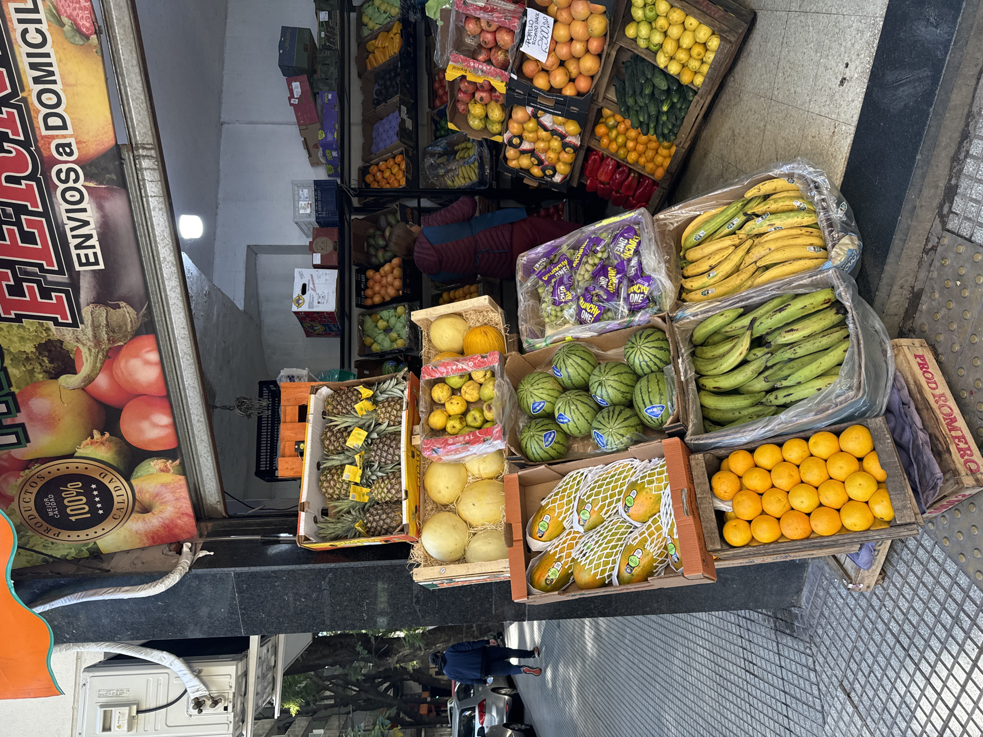 This is an outdoor scene displaying a fruit and vegetable stand in what appears to be a bustling city environment, most likely in Buenos Aires, Argentina, given the Spanish signage. The stand is overflowing with a variety of fresh produce, primarily fruits, neatly arranged in wooden crates and boxes. There are pineapples, melons, watermelons, papayas, bananas, oranges, and other fruits such as apples and grapes visible. These fruits are stacked on and around the ground in the doorway of what appears to be a storefront.
A sign for the store is visible, featuring the phrase "ENVIOS a DOMICIL", indicating delivery services. The items are arranged to entice passing customers.
A person is partially visible, standing near the curb, and a white car is parked on the street behind the store. The ground is covered in grey tiles.
