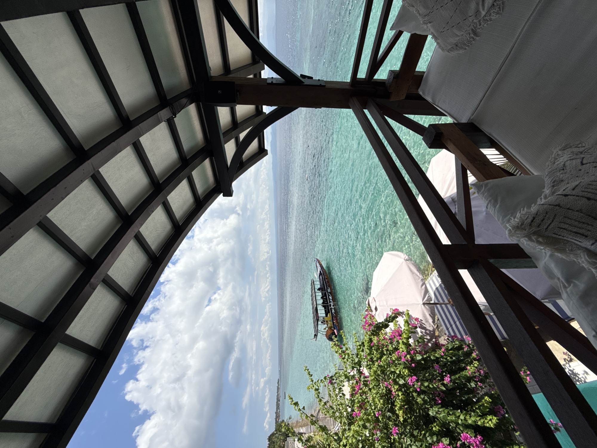 The photograph captures a coastal scene from Nusa Penida, Indonesia. The image is framed by a dark wooden structure, possibly part of a beachside gazebo or pergola, which contrasts with the bright seascape. A longtail boat is anchored in the clear, turquoise water. Green vegetation with bright pink flowers, likely bougainvillea, cascades along the bottom right portion of the frame. In the distance, a coastline and hints of buildings can be seen. The sky is partially covered with clouds. Adjacent to the wooden structure are white lounge cushions and an umbrella.
