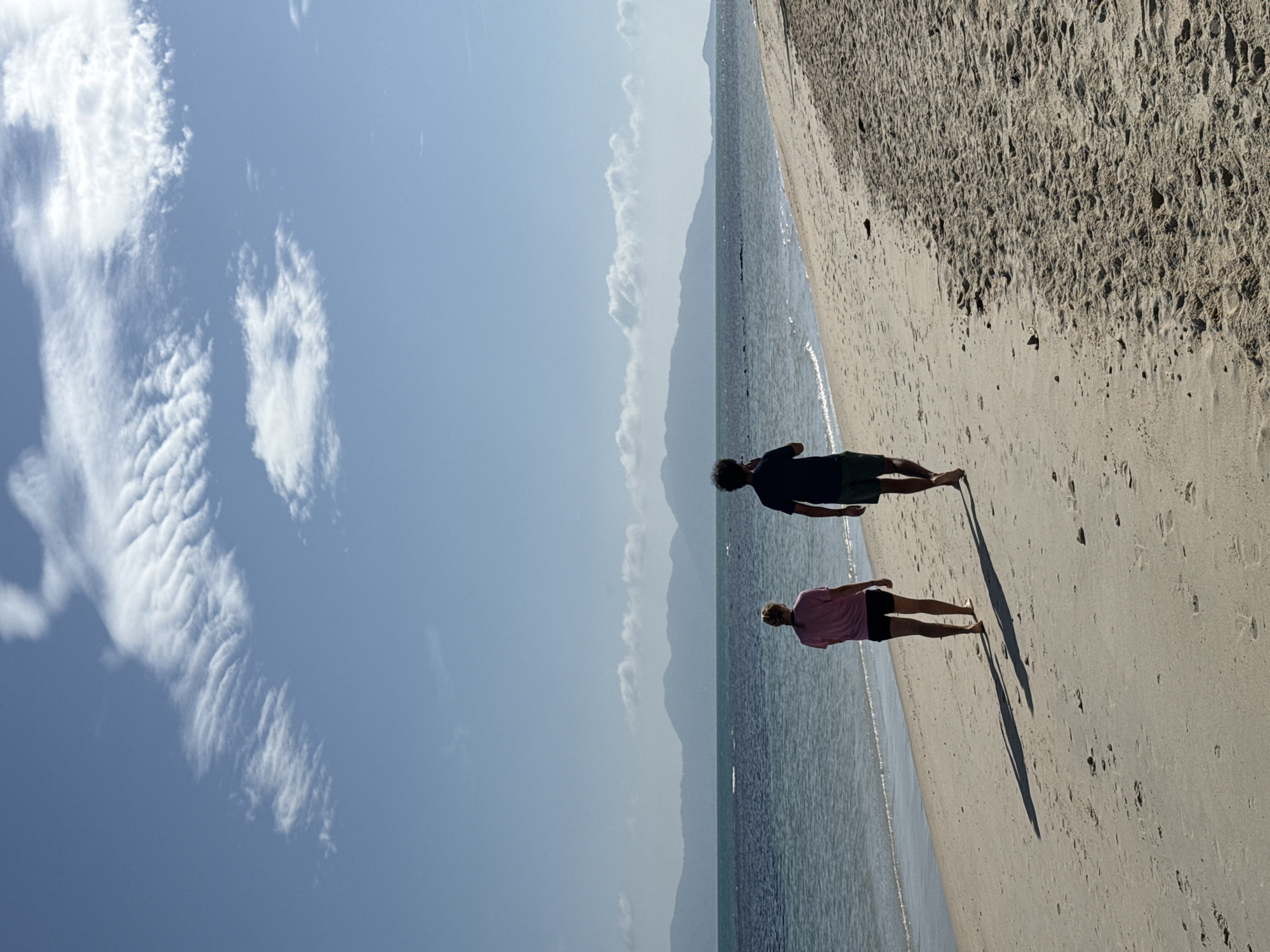 Two individuals, a woman and a man, walk barefoot along a sandy beach in the Ko Samui District of Thailand. The woman, on the left, wears a pink t-shirt and black shorts, her hair in a braid. The man, on the right, wears a dark t-shirt and green shorts, and appears to be holding a phone to his ear. They walk away from the camera, casting long shadows on the sand. The calm, blue ocean borders the beach to their left, and in the distance, a range of hazy mountains forms the horizon. The sky above is a clear blue, with a few wispy white clouds scattered across it. The overall scene suggests a peaceful, sunny day.