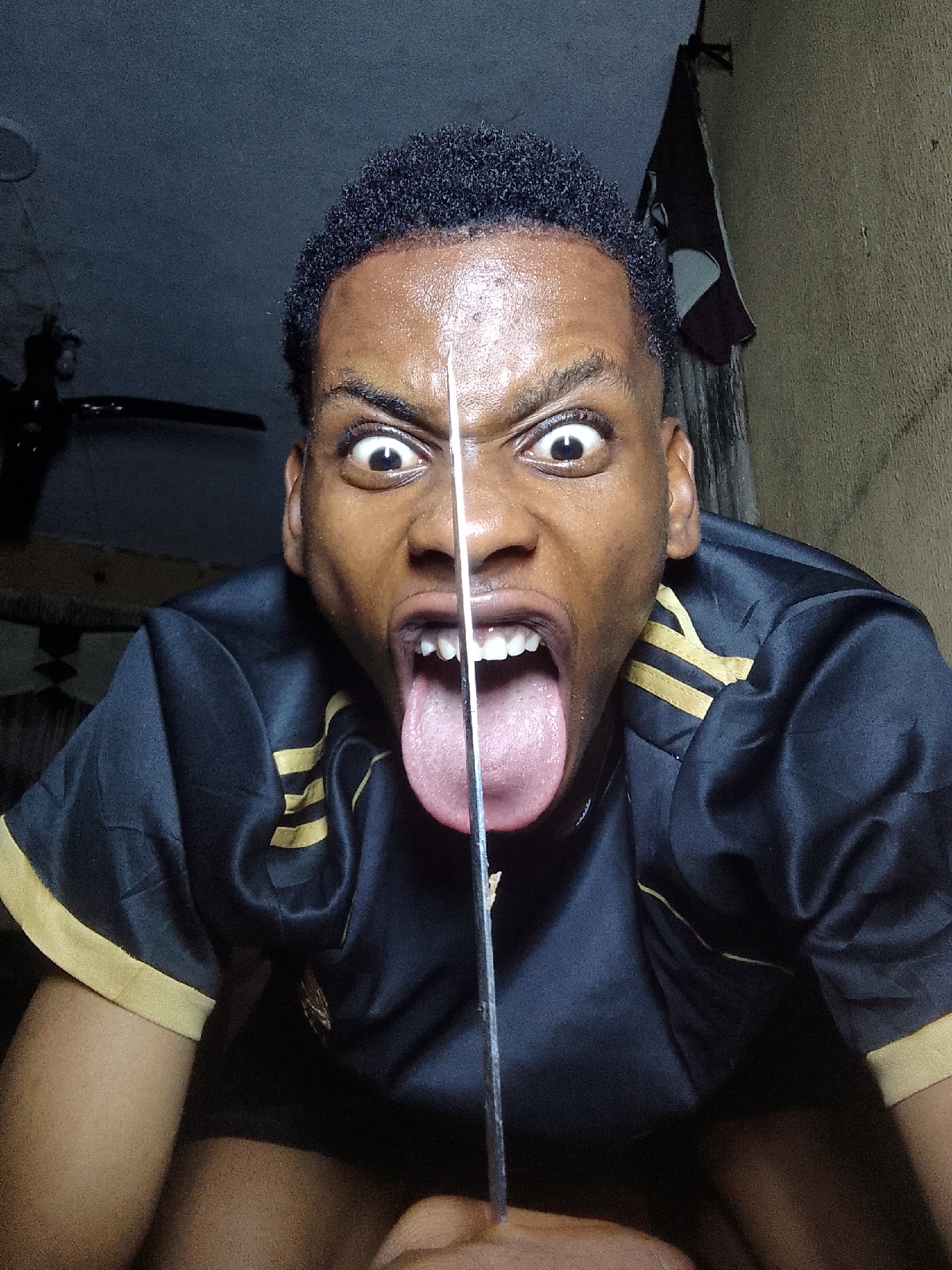 The image shows a dark-skinned person with short, dark hair, making a distorted facial expression with wide eyes and an open mouth with tongue extended. A metallic object, likely a knife, is positioned vertically, bisecting the face from forehead to tongue. The person is wearing a black shirt with gold stripes on the sleeves. The background is a dimly lit room with a ceiling fan visible in the upper left and a cloth hanging in the upper right. The location is Unknown, Unknown.