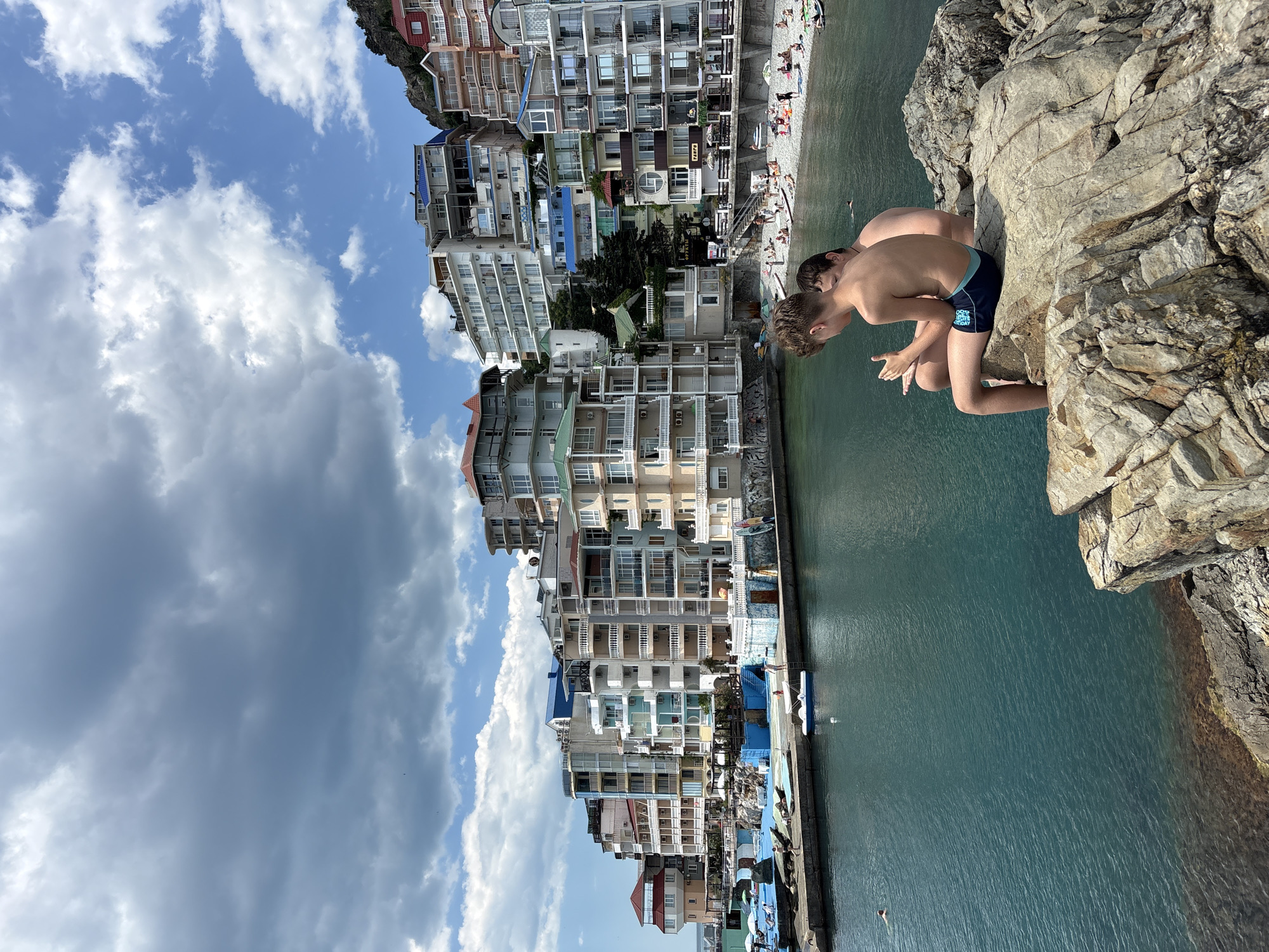 The image depicts a coastal scene in Kiparisnoe, Ukraine, characterized by a dense collection of multi-story buildings lining a waterfront. In the foreground, two young boys are seated on large rocks, facing away from the camera and towards the water. One boy is wearing dark blue swim trunks, and the other is out of focus behind him. The water is a vibrant turquoise color, and the shoreline is lined with a pebbly beach populated by many people sunbathing and swimming. A small harbor area with several boats is visible to the left, and further along the coast, more buildings are clustered on the hillside. The sky is partly cloudy with dramatic, dark clouds interspersed with patches of blue.